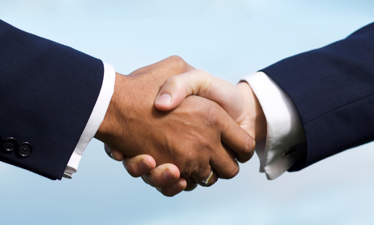 Two men in suits shake hands against a blue sky