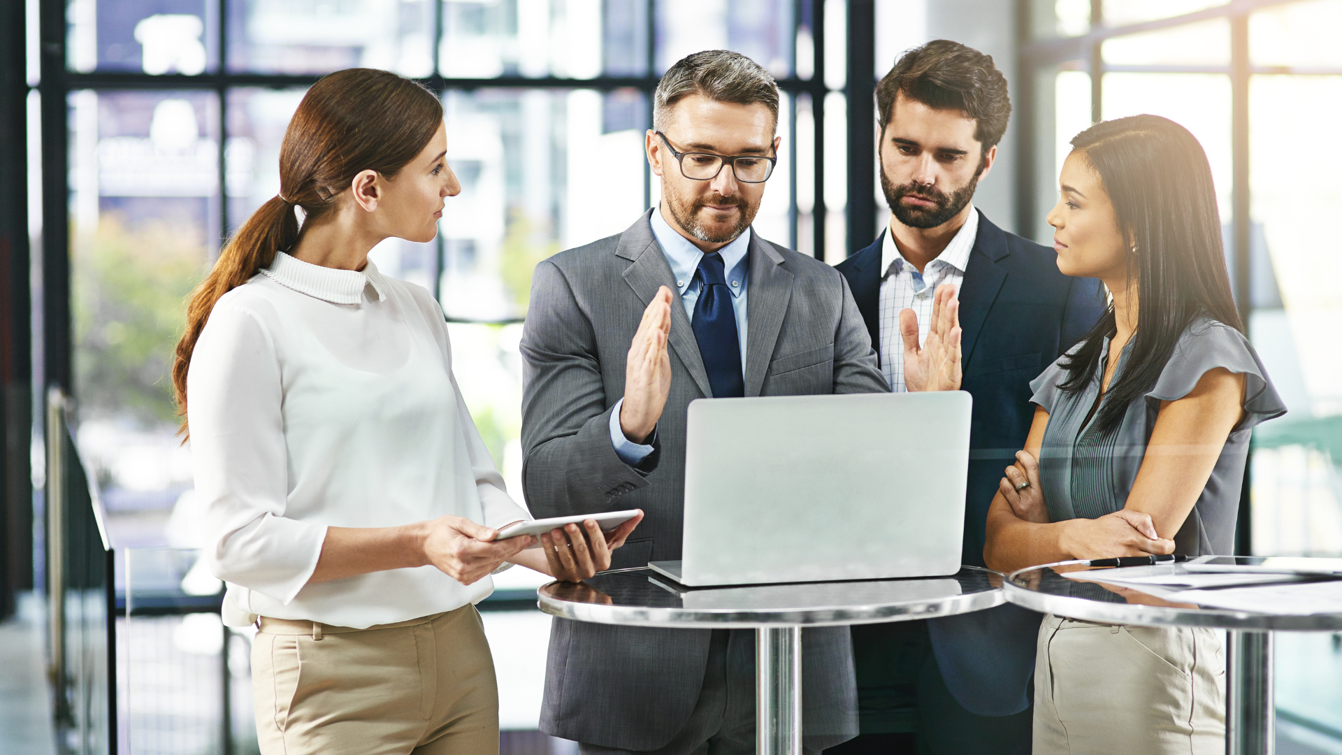 Four businesspeople in suits at a laptop, gesturing, discussing in an office setting.