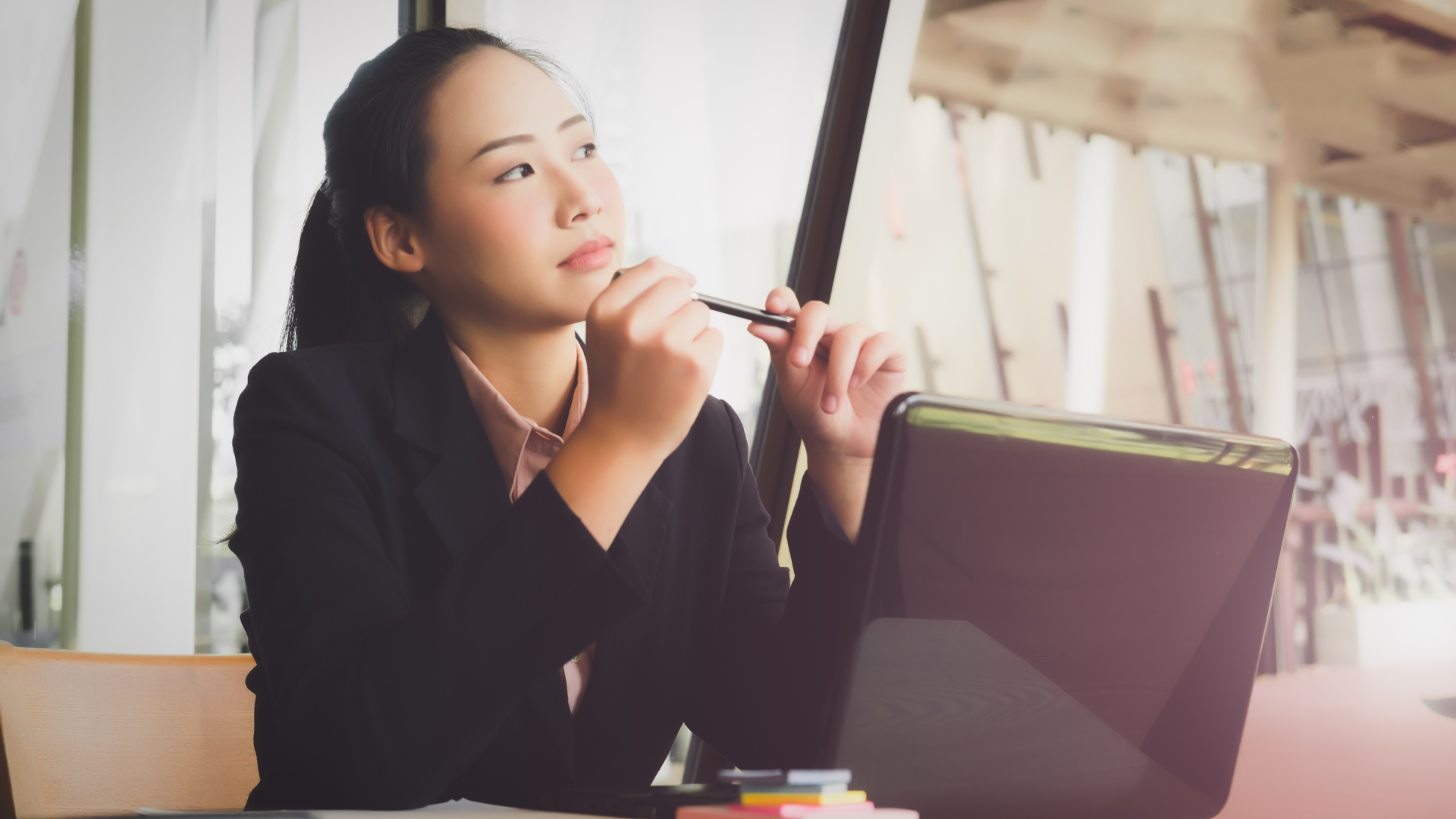 A woman is sitting at a desk with a laptop and a pen in her hand.
