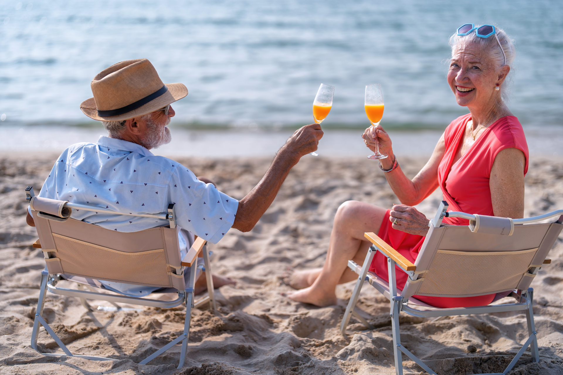 An elderly couple is sitting on the beach toasting with wine glasses.