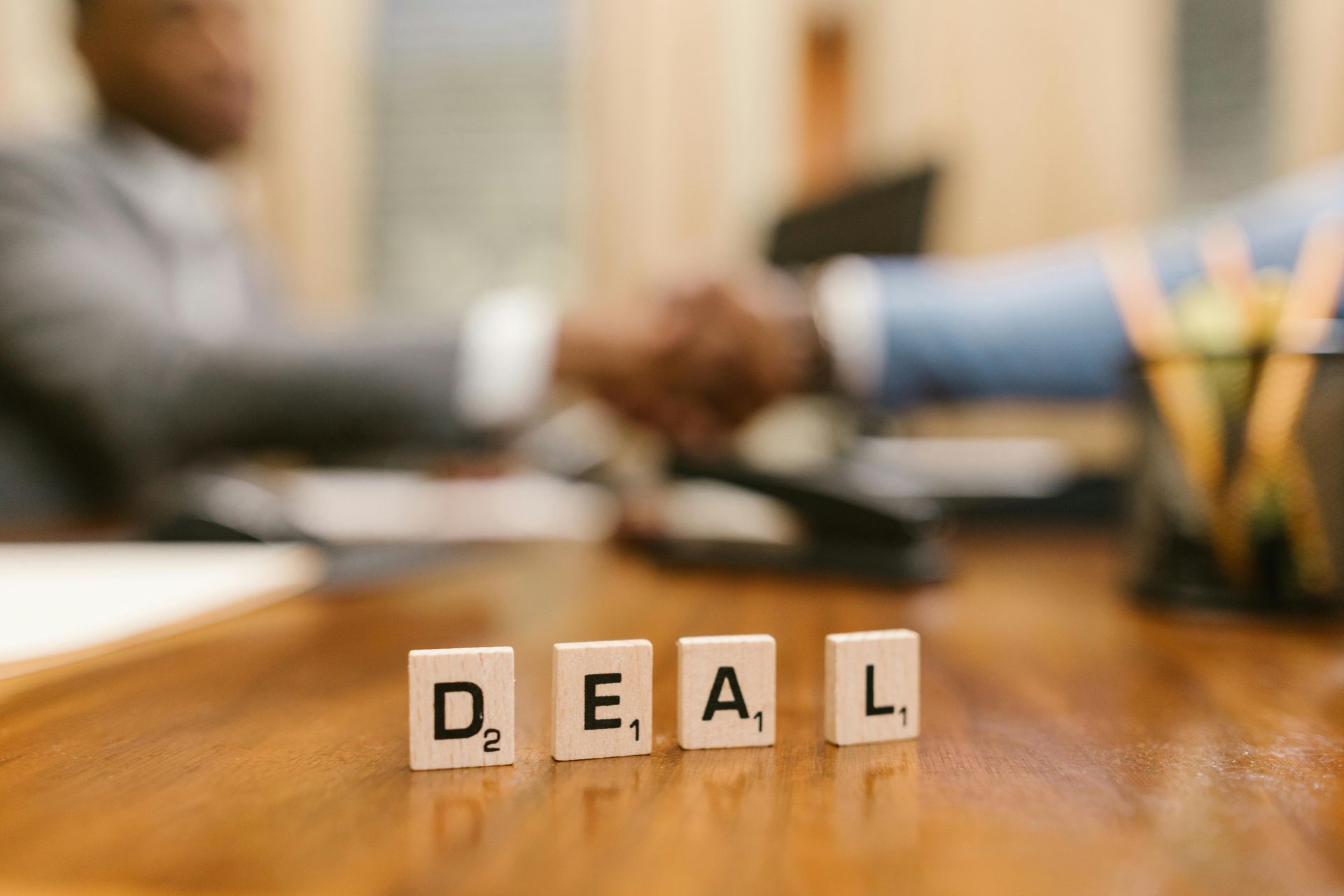 A man is shaking hands with another man behind a table with scrabble tiles that spell out the word deal.