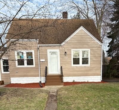 A tan-sided house with a dark roof and a small front porch, surrounded by bare trees and grass under a blue sky.