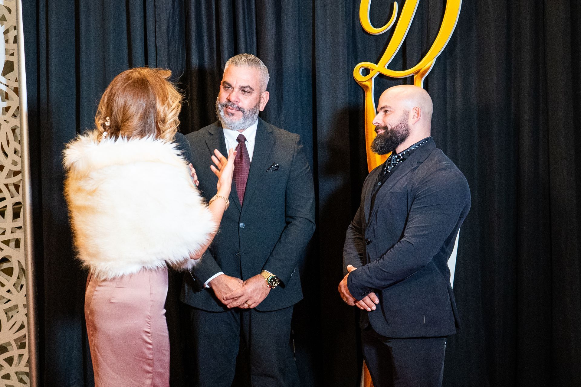 Three people in formal attire speaking on a stage with black curtains and a gold logo backdrop.