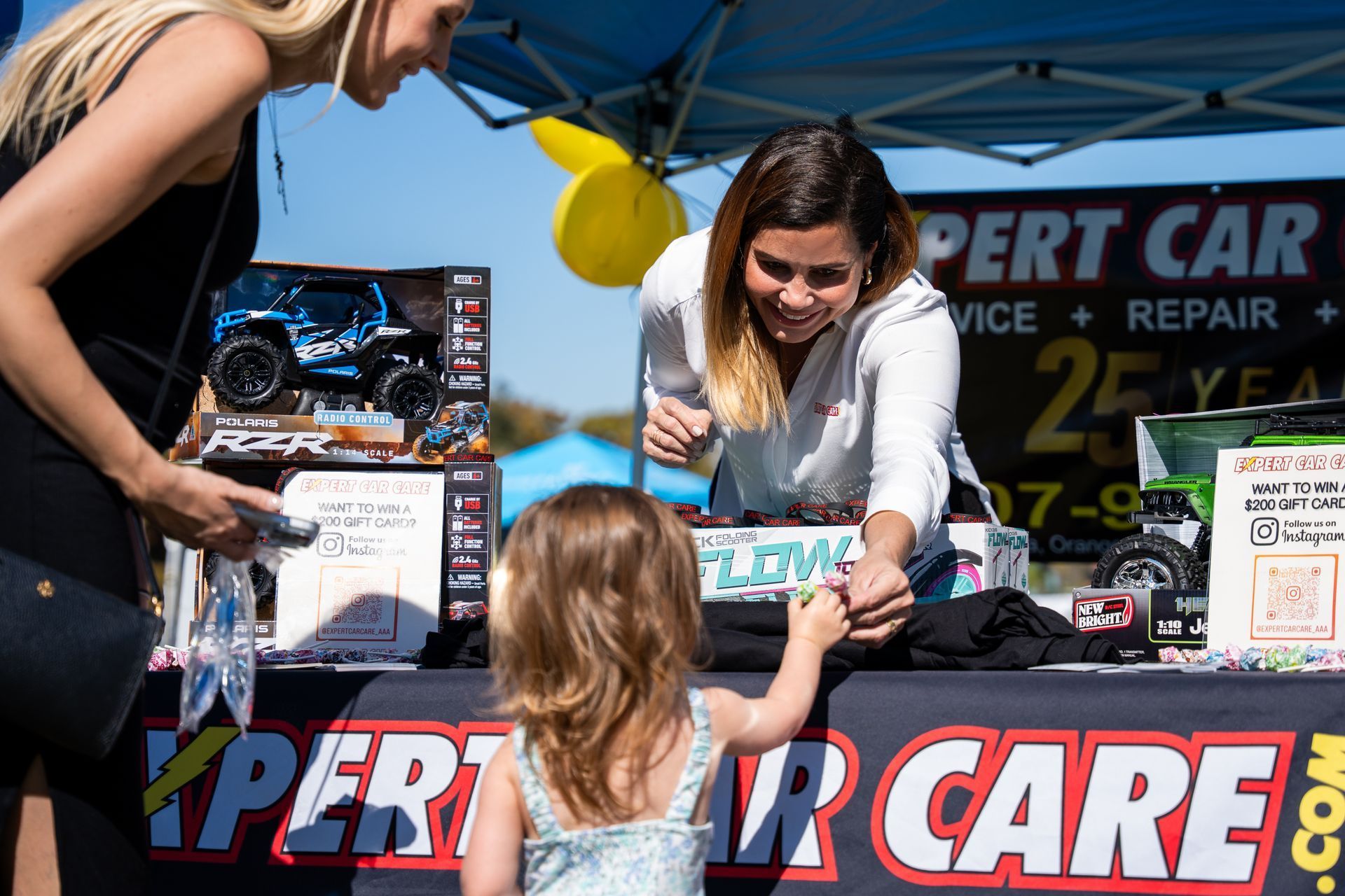 Two women at an Expert Car Care booth hand a sign to a child at an outdoor event.