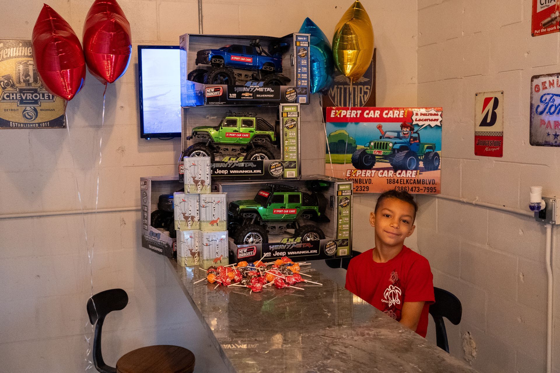 Boy seated behind a table stacked with toy car boxes and balloons in a decorated room