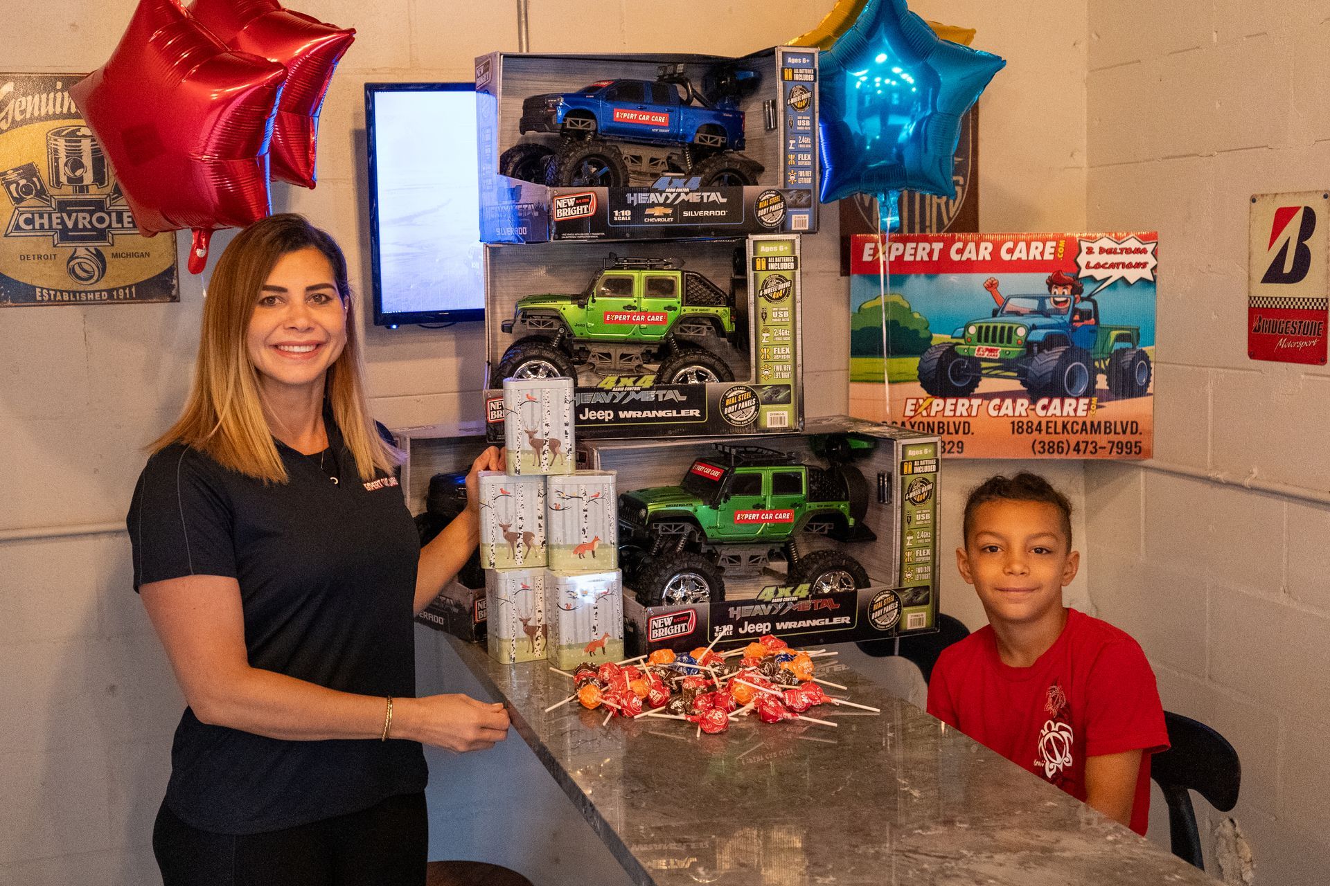 Woman and boy beside a large stack of boxed toy cars and gift packages at a counter with balloons