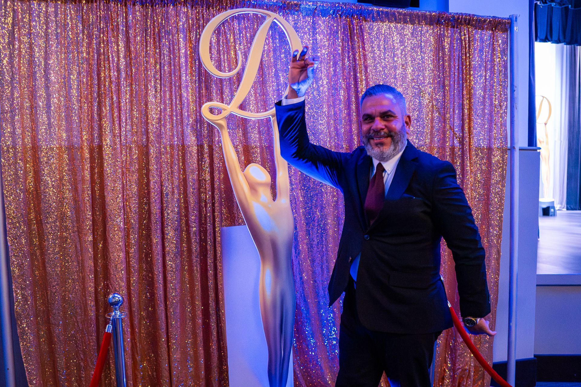 Man in a dark suit posing beside a gold sequin backdrop and white mannequin at a formal event