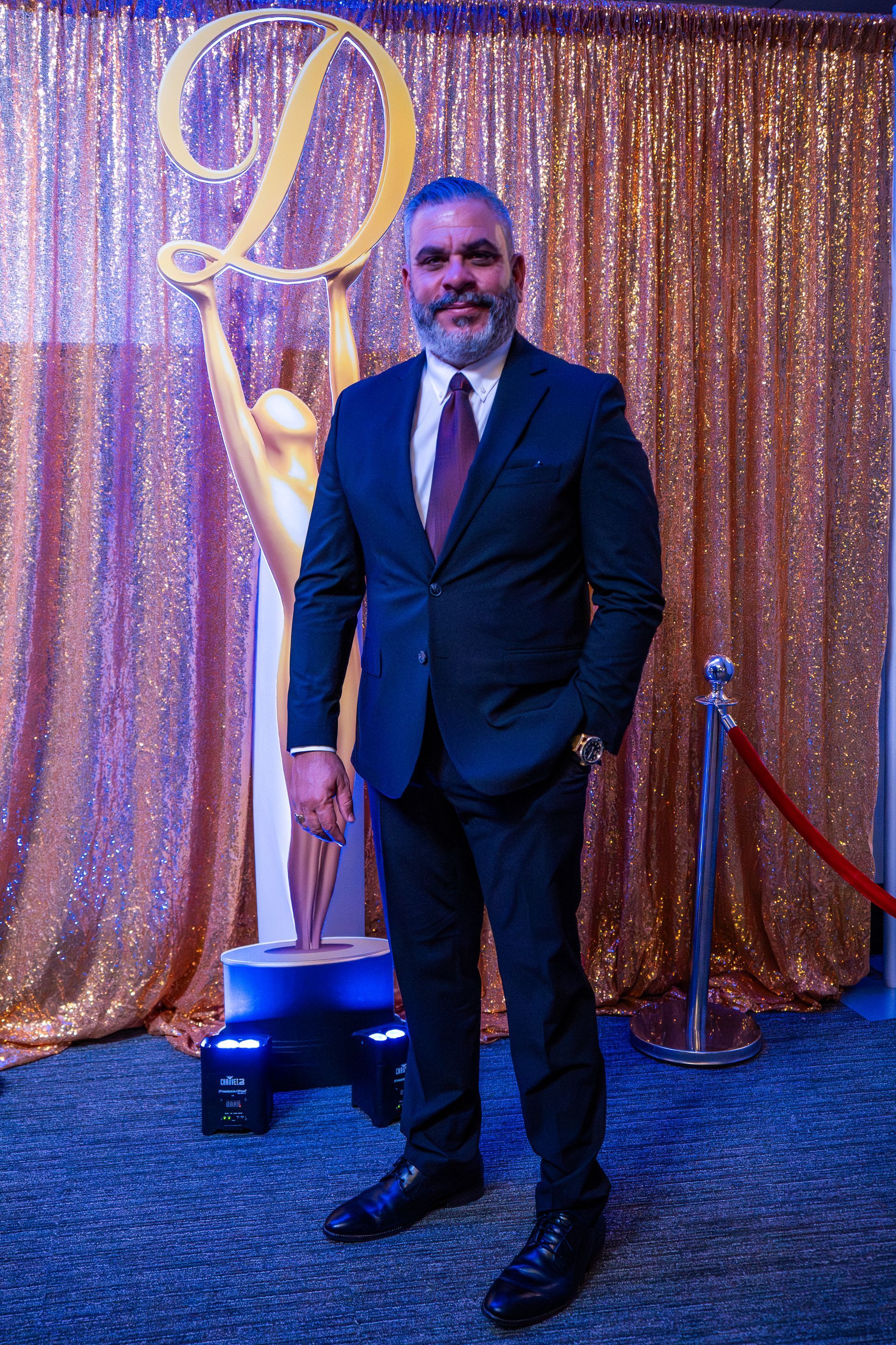 Man in a dark suit posing in front of a gold sequin backdrop and award statue at an event