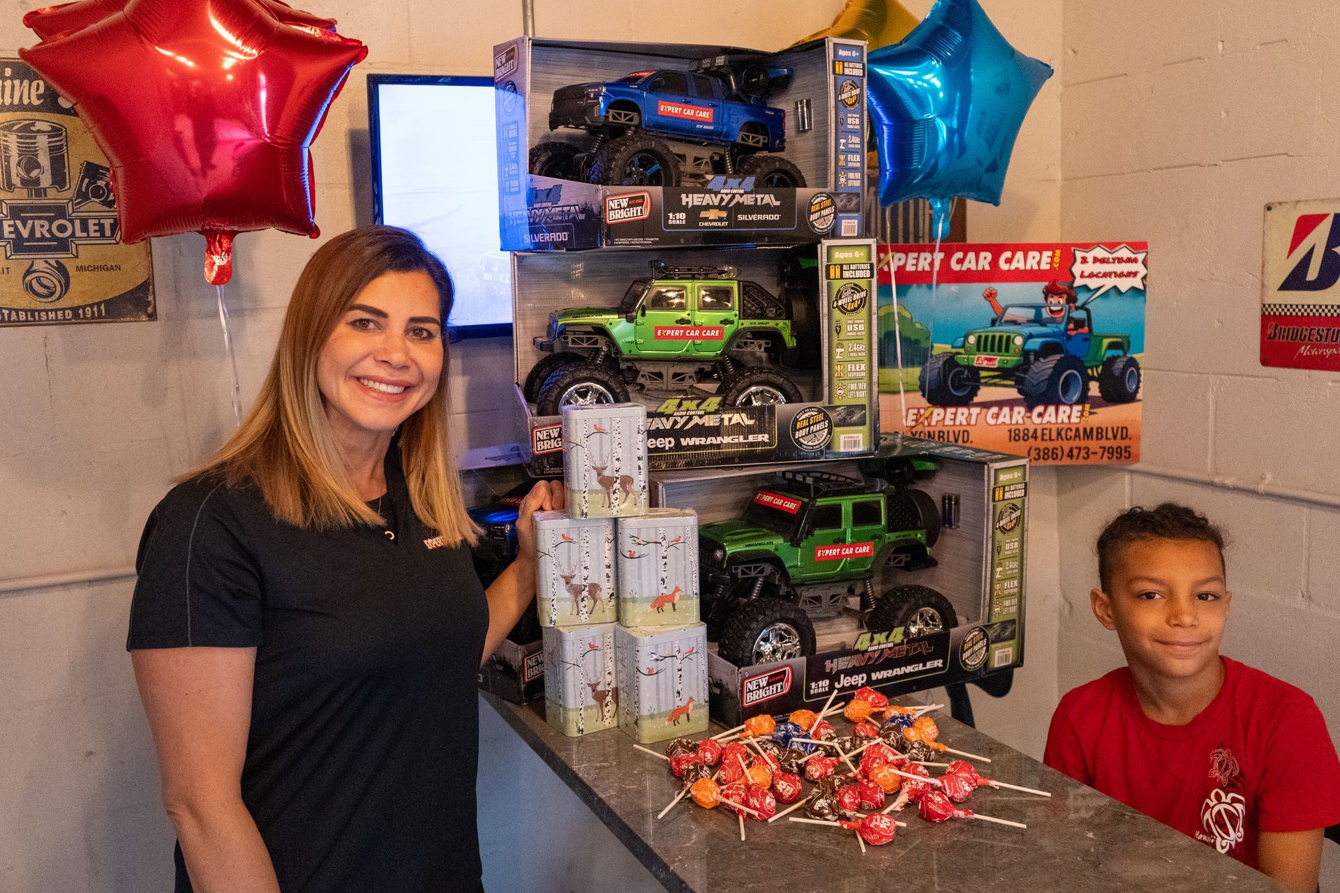 Woman and boy beside stacked toy car boxes, balloons, and wrapped gifts in a store display.