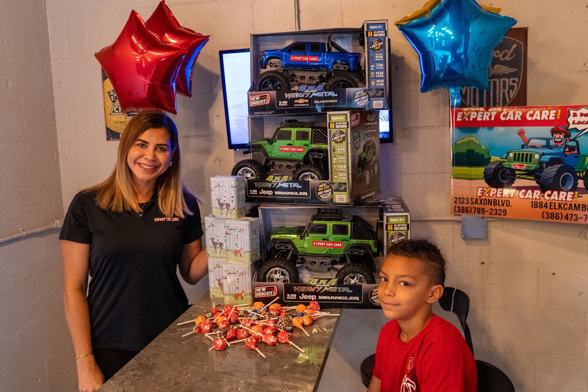 Children beside a large toy truck display with red and blue star balloons and a pile of wrapped treats