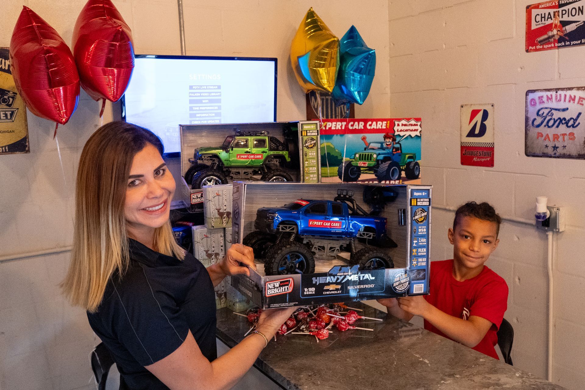 Woman and child holding a boxed toy car set in a decorated room with balloons and a TV in the background