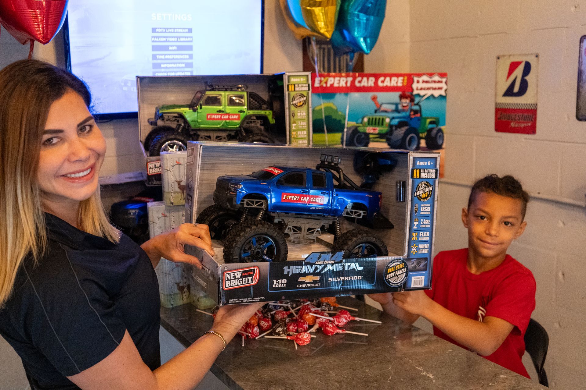 Woman and child holding a boxed remote-control car with balloons and small toy cars on a table