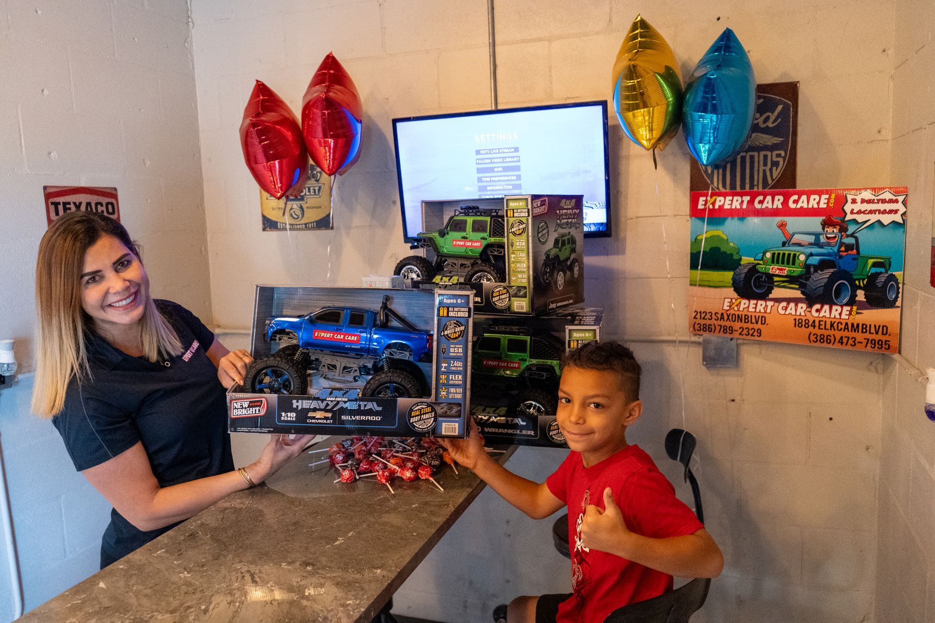 Woman and boy posing by colorful toy car displays in a room with a wall-mounted screen
