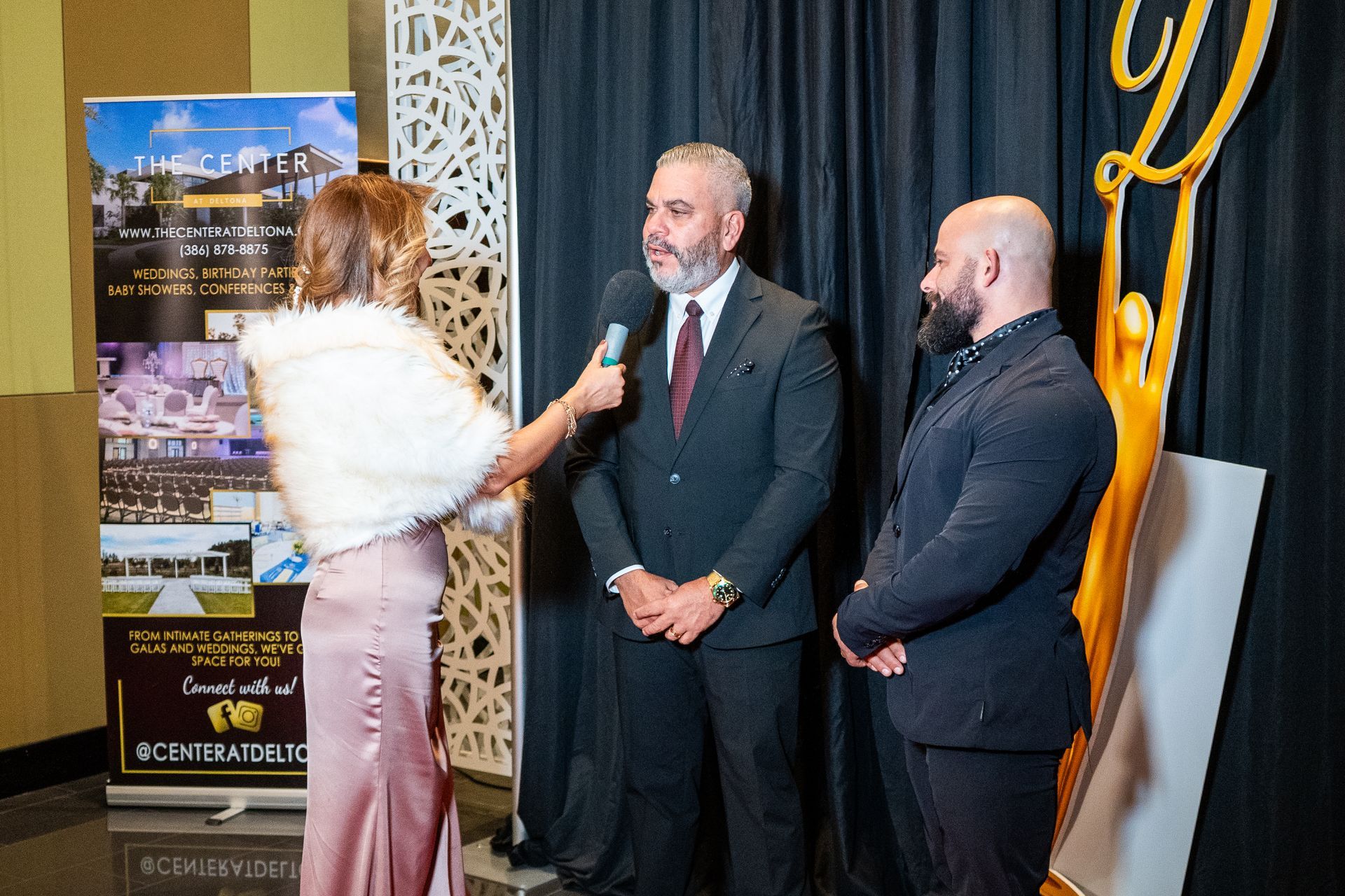 Woman interviewing two men at an indoor event beside a poster display