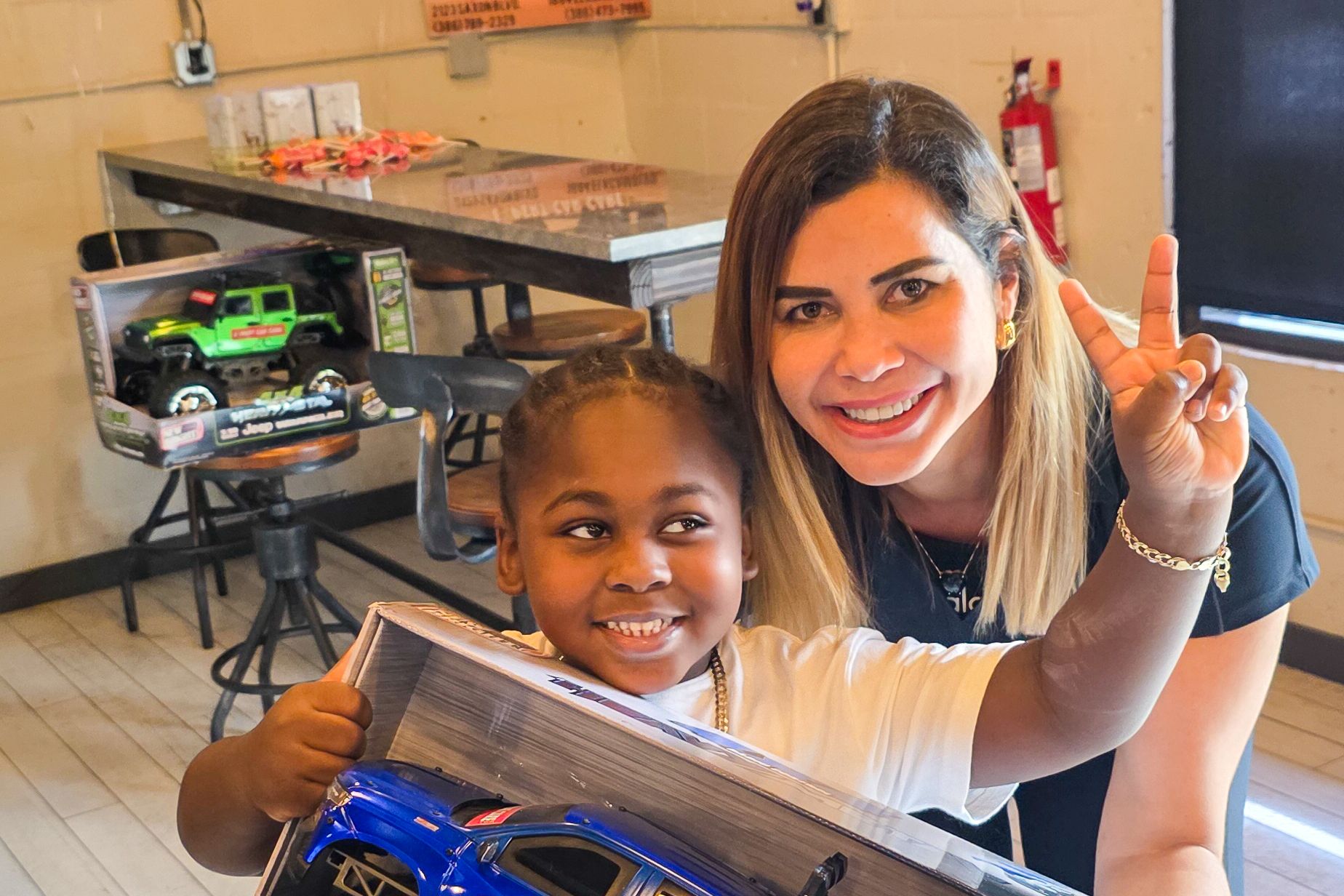 Two smiling people posing with toy cars in a workshop, one making a peace sign.