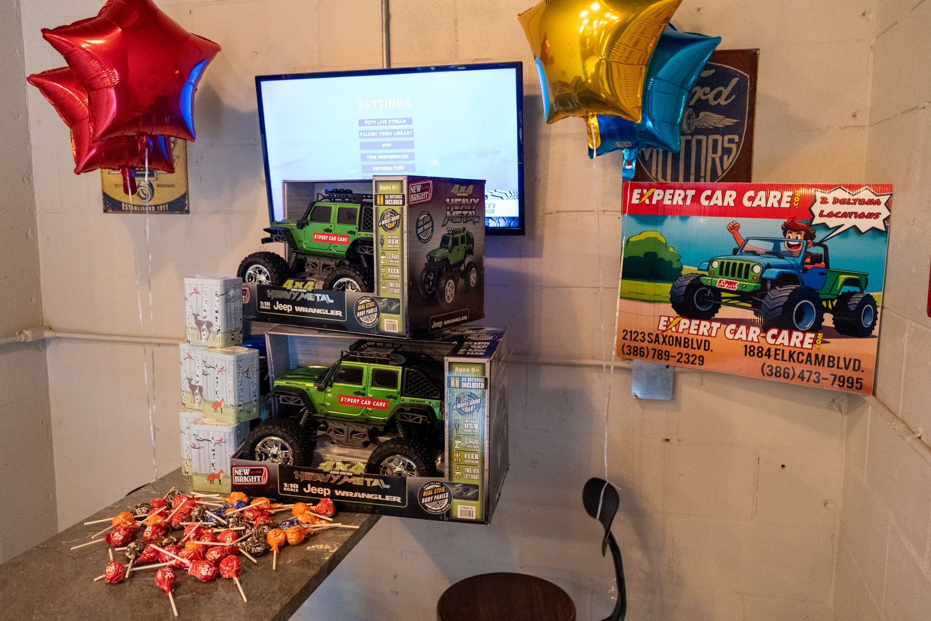 Toy monster trucks displayed on a table with star balloons and a TV in the background