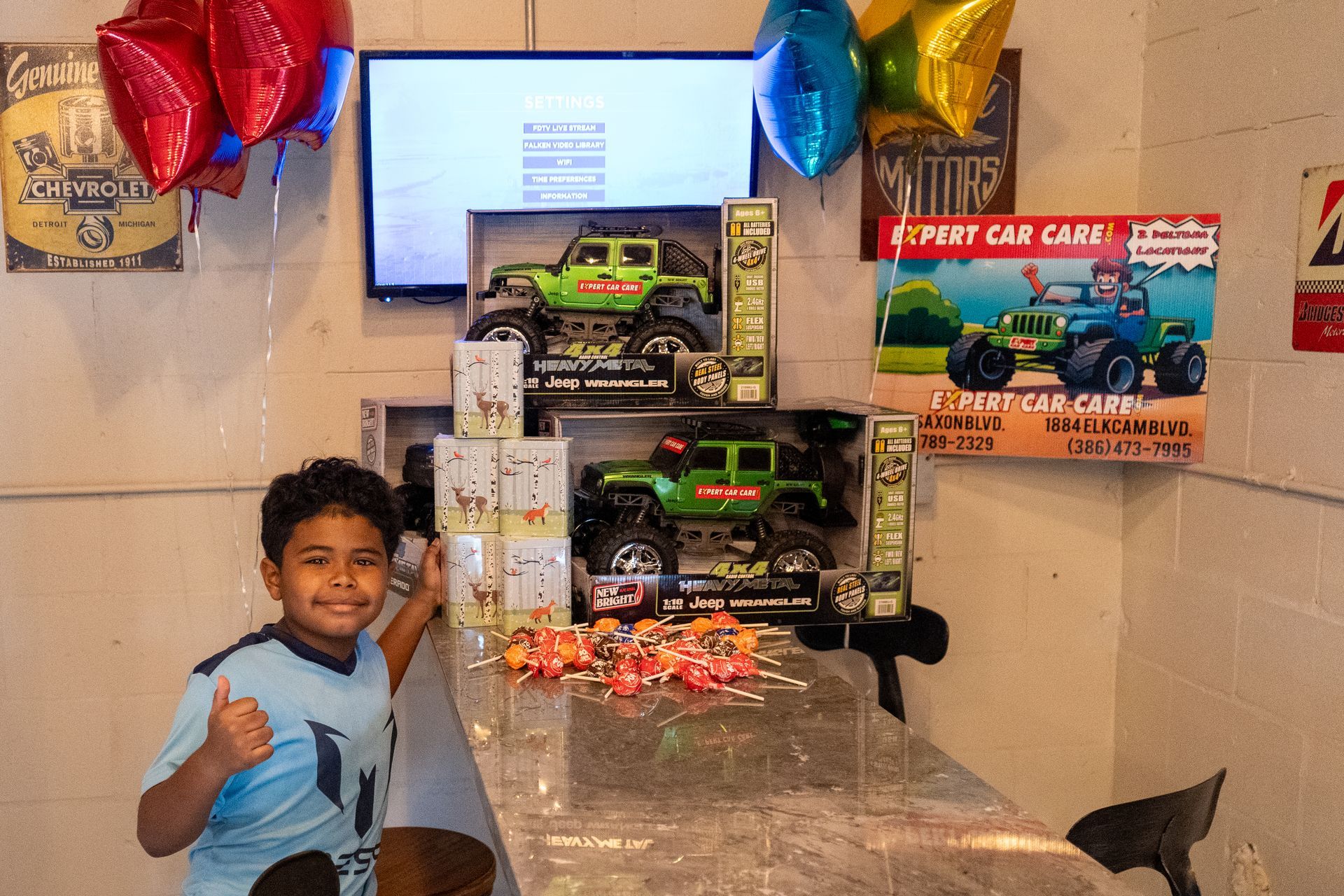 Boy smiling beside stacked toy car boxes and balloons on a table.