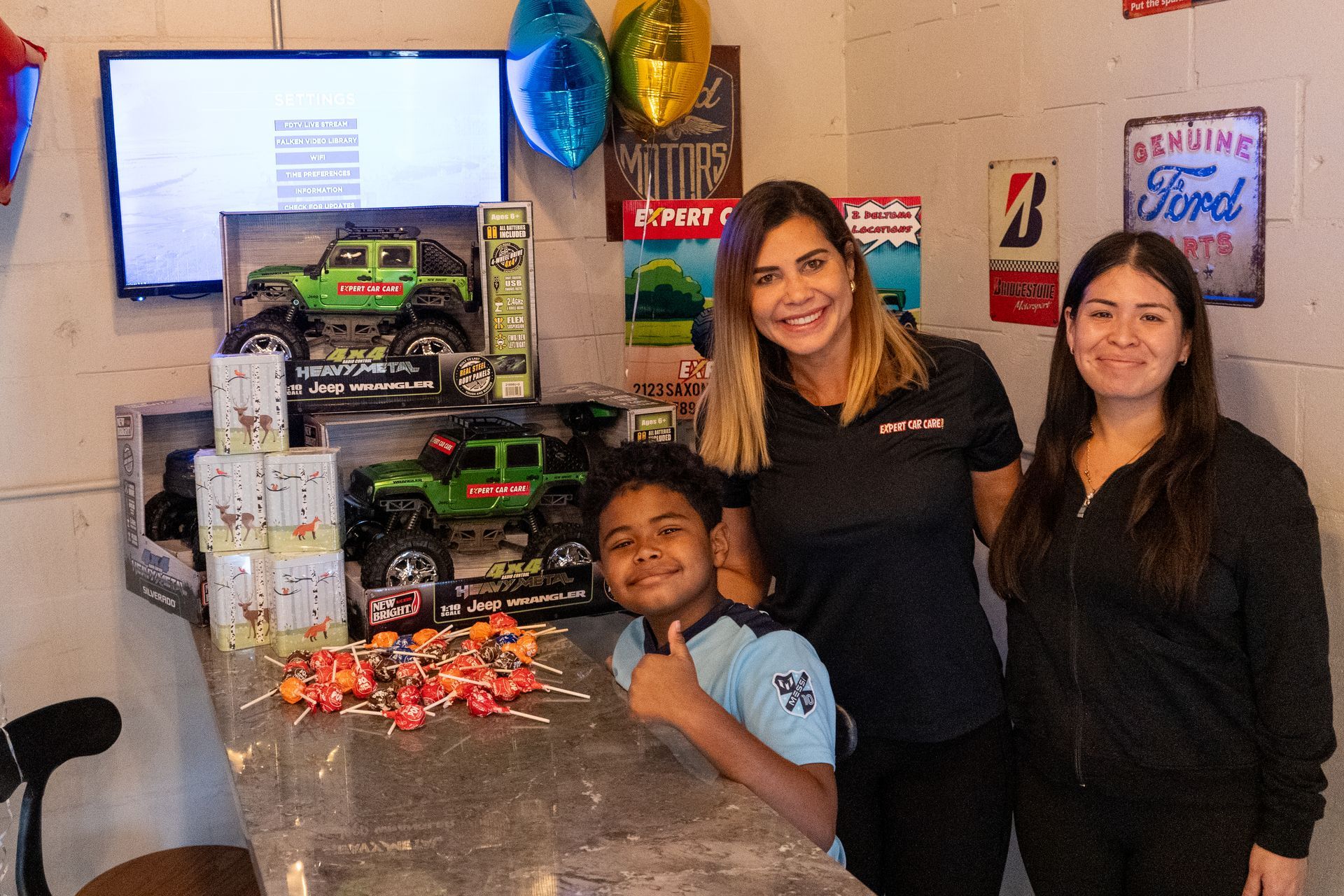 Three people standing at a counter with toy cars, balloons, and a decorated display in the background