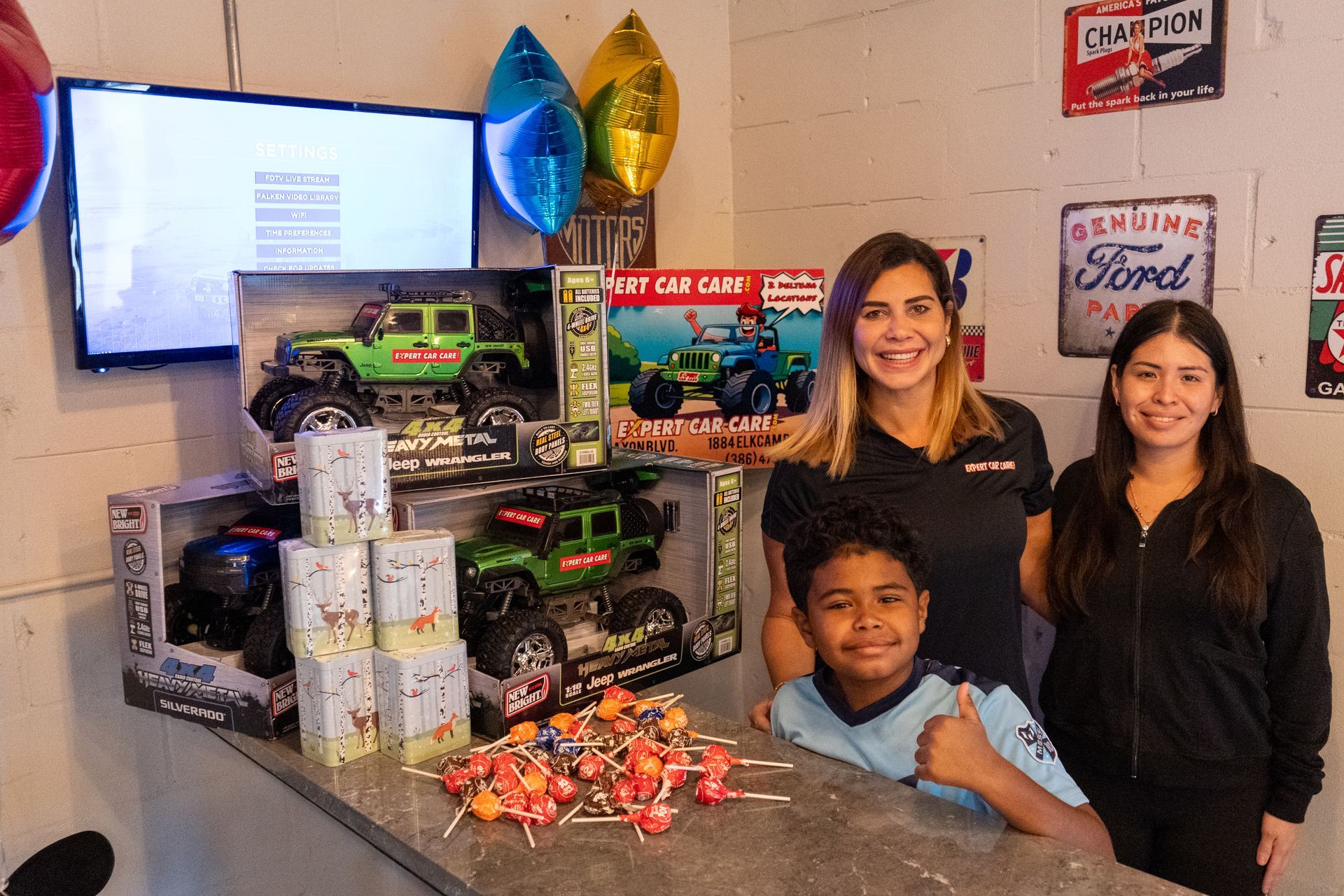 Three people posing beside a toy display with green trucks and balloons in a decorated room