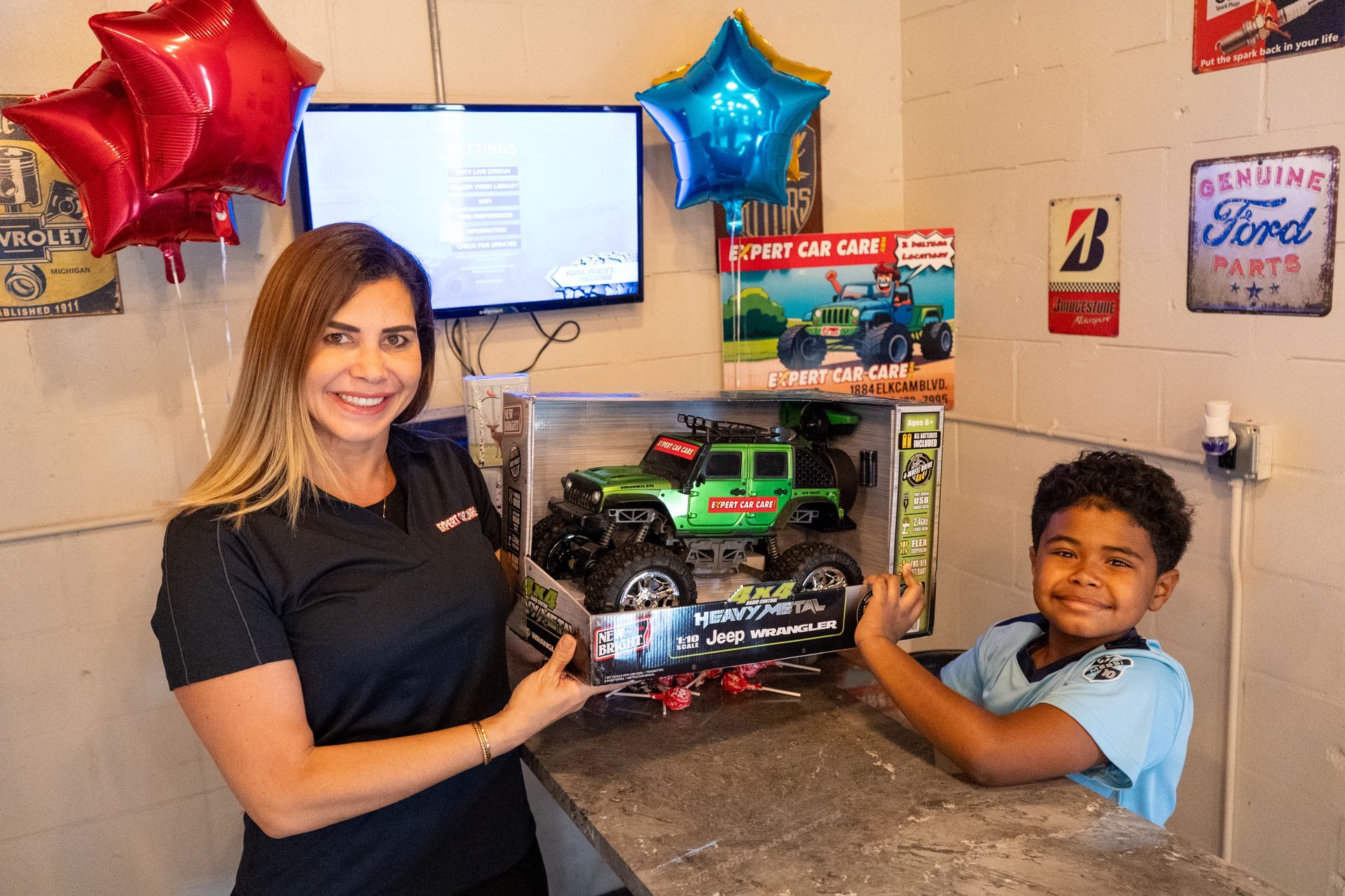 Woman and child holding a boxed toy truck in a decorated room with balloons and a wall-mounted screen.