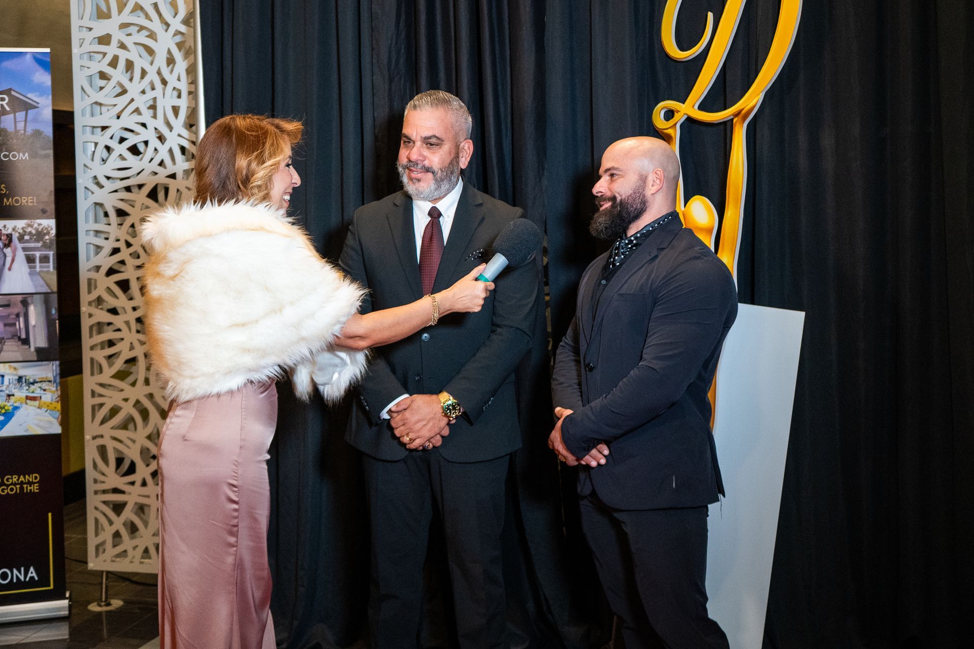 Three people at a formal event, with a woman holding a microphone toward two men in suits.