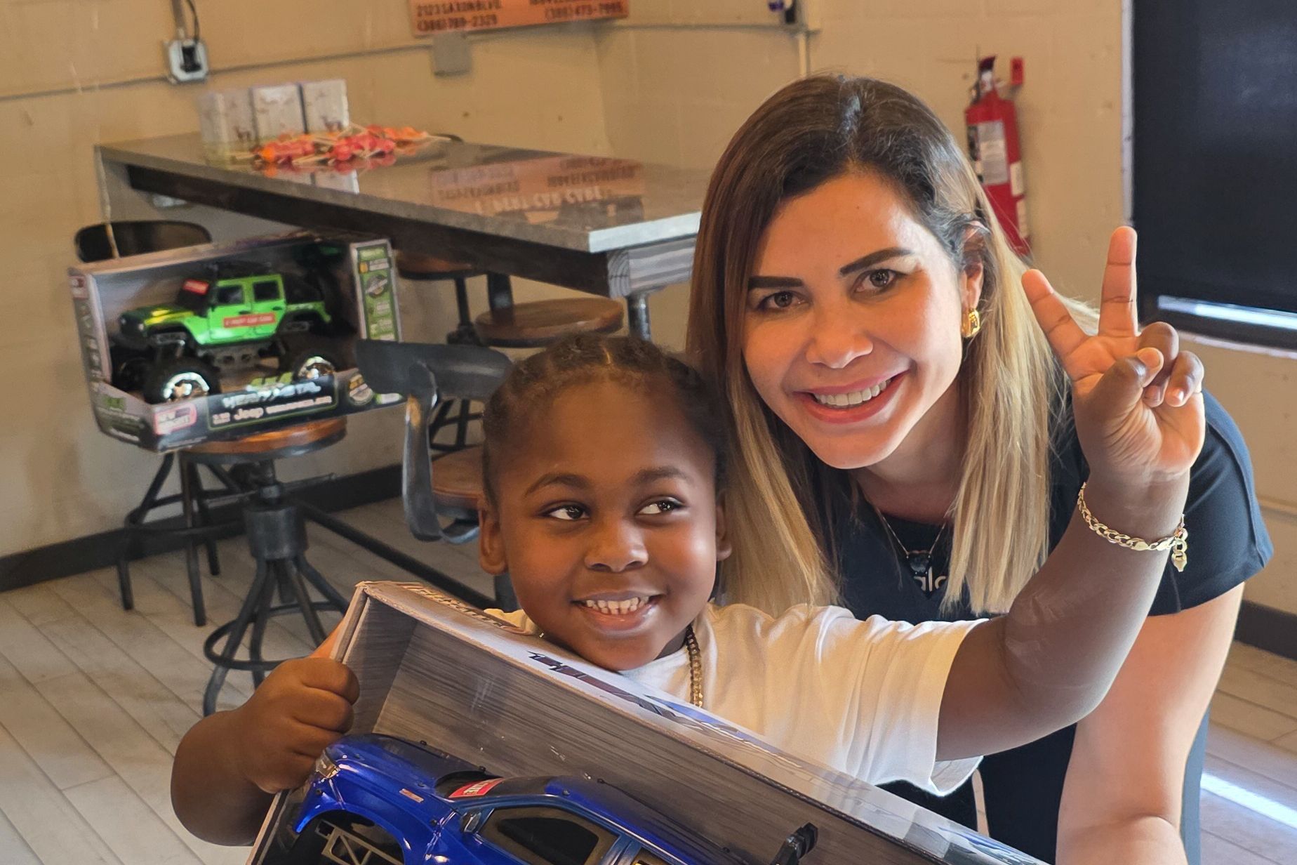 Two smiling people holding toy cars in a workshop, one flashing a peace sign.