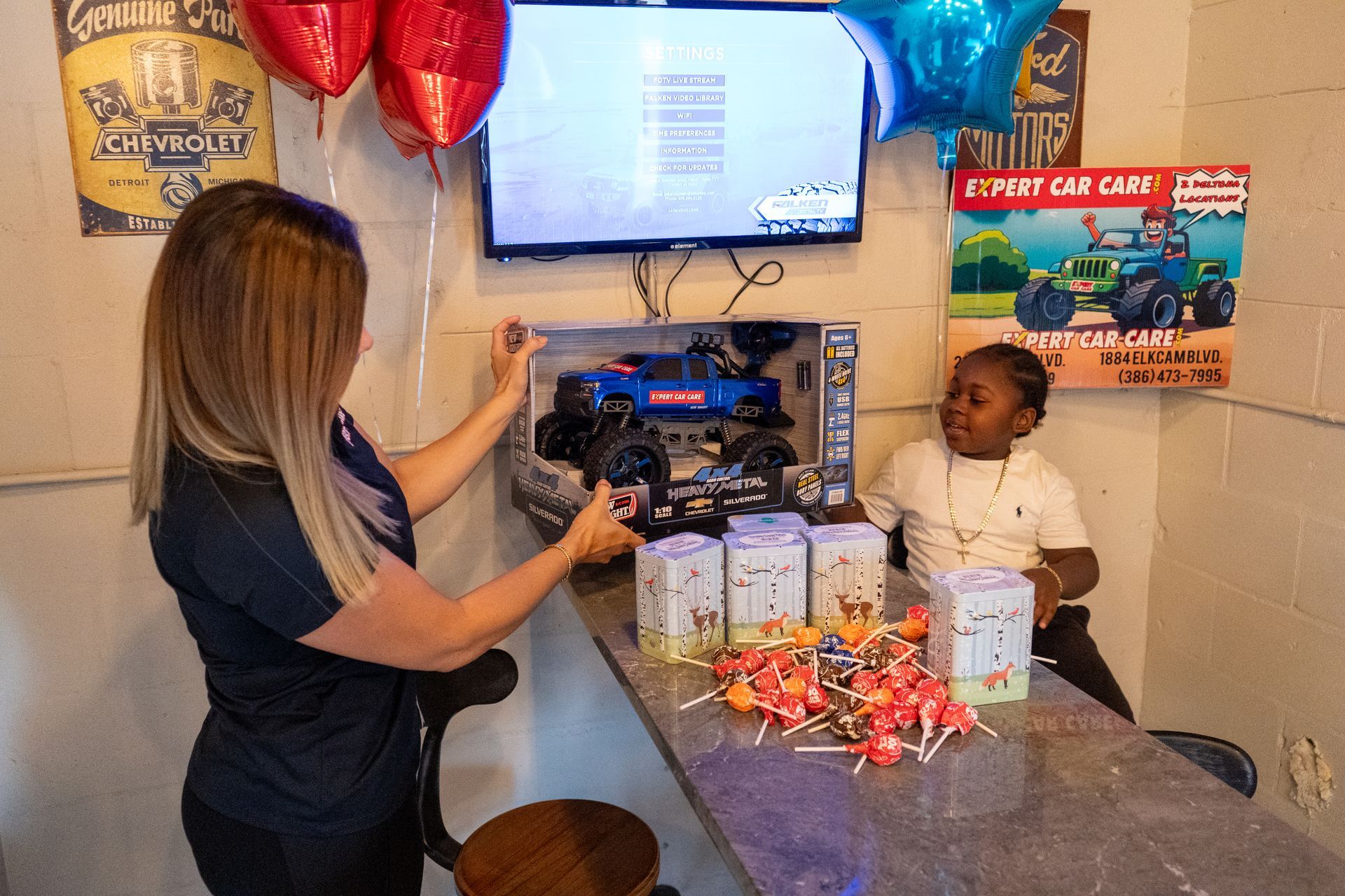 Woman and child at a birthday party table with gifts, balloons, and decorations under a wall-mounted TV