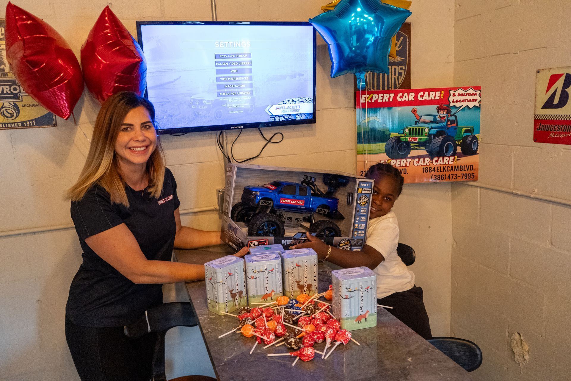 Woman and child at a table with gift bags, balloons, and a wall-mounted screen in a classroom or office setting