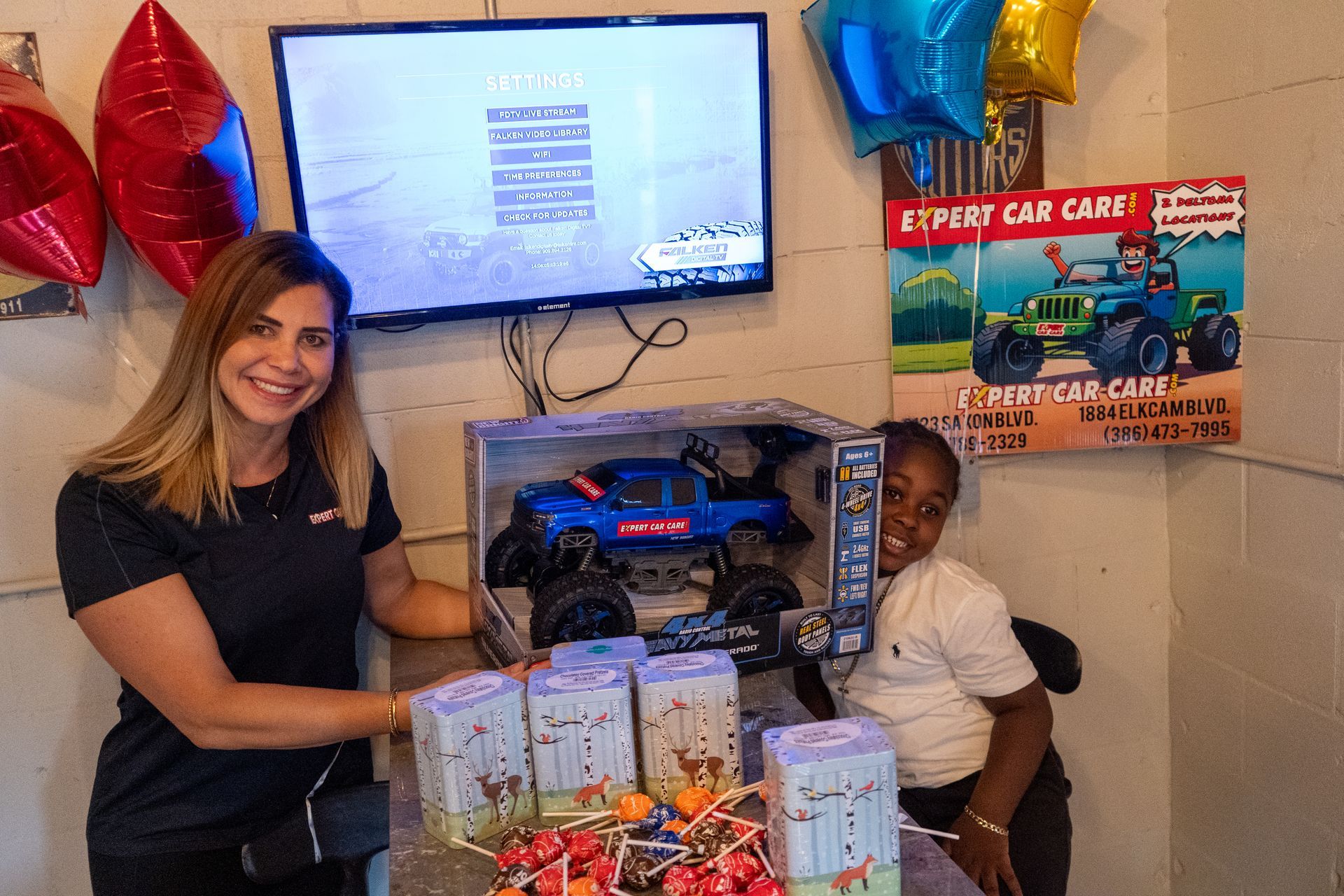 Woman smiling beside toy car gift display with balloons and a TV in the background