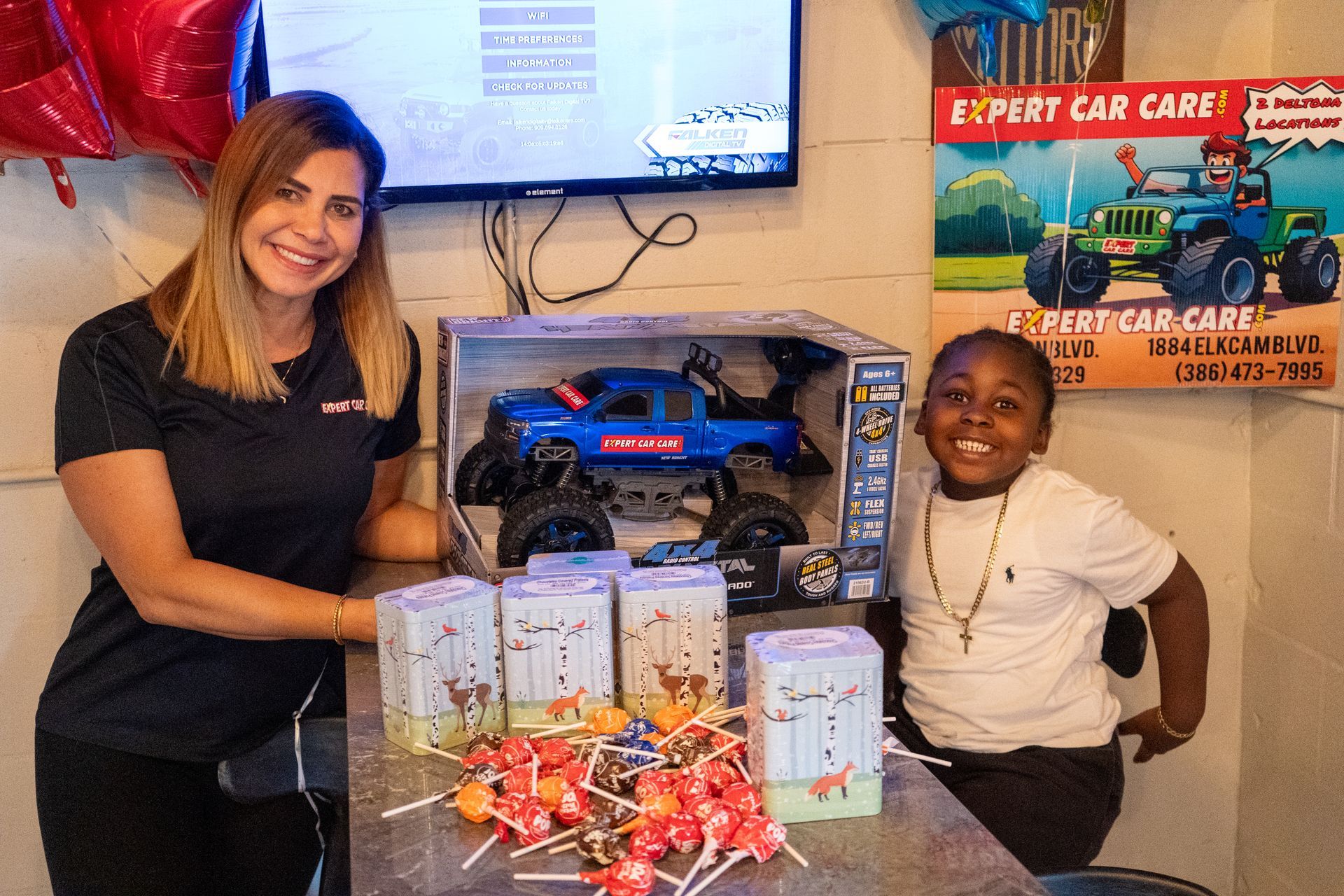 Two people pose beside a boxed toy truck and wrapped gifts at a car care display.