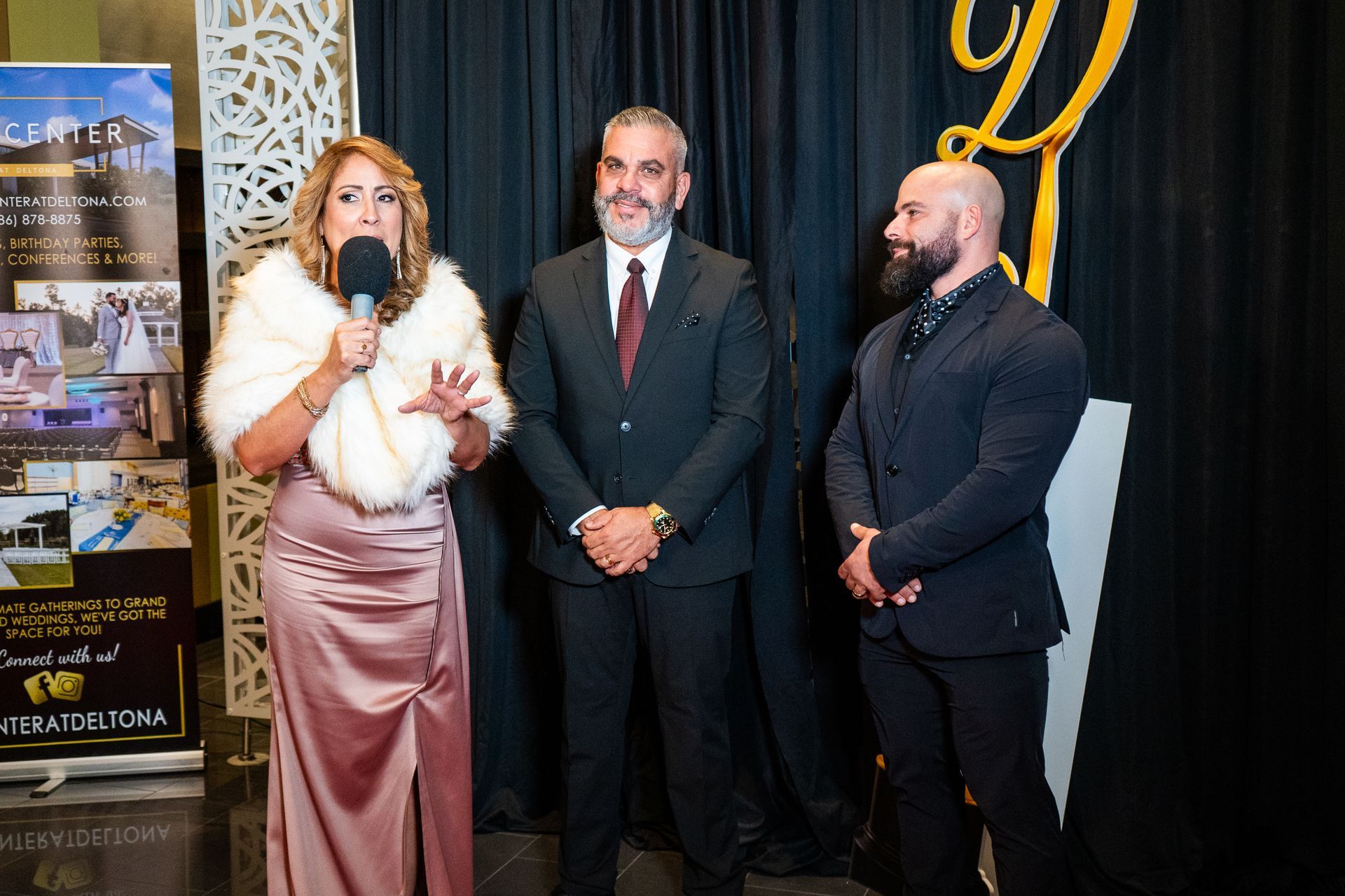 Woman speaking into microphone beside two men at a formal event backdrop