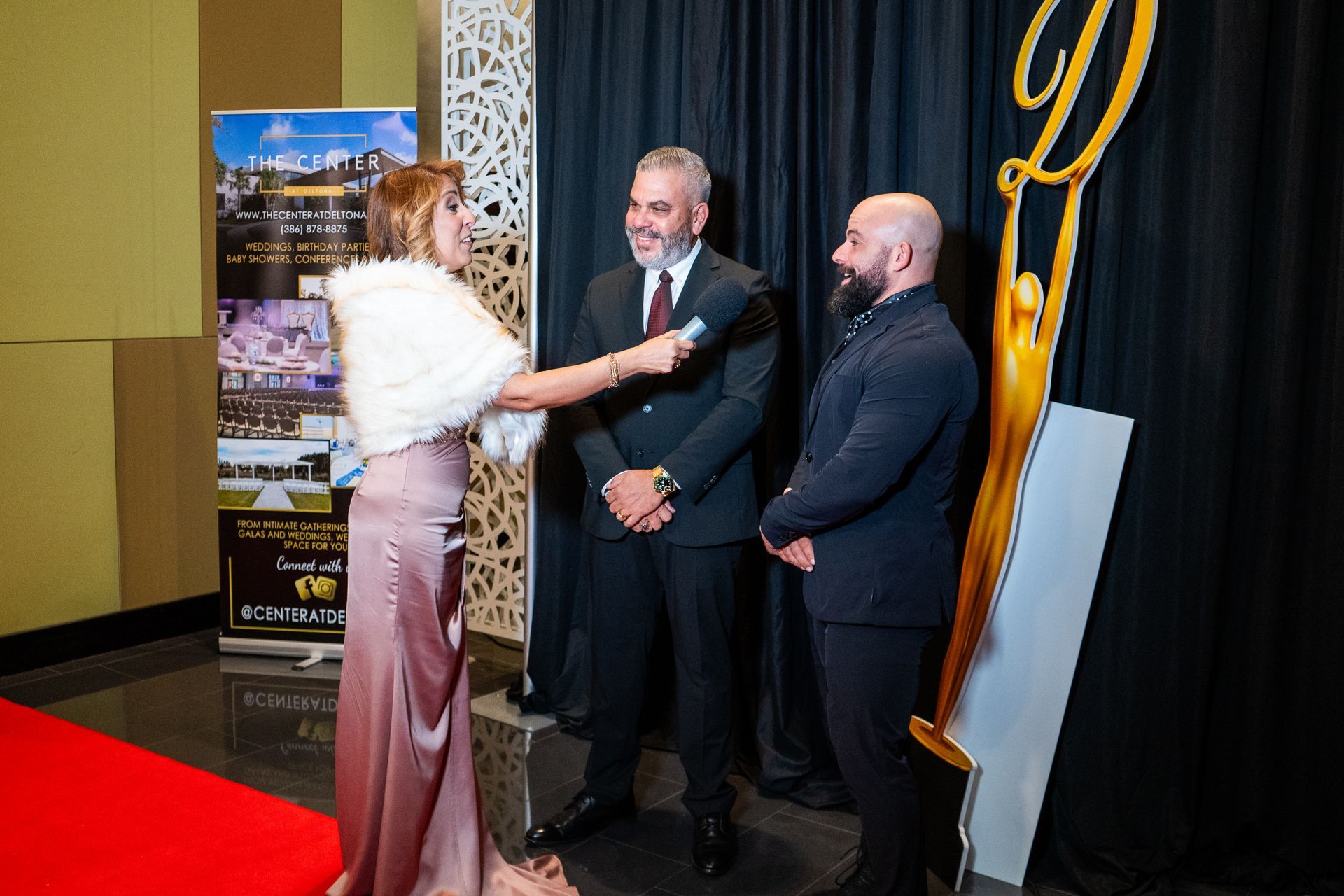 Woman in a white fur wrap greets two men in suits at an indoor event backdrop