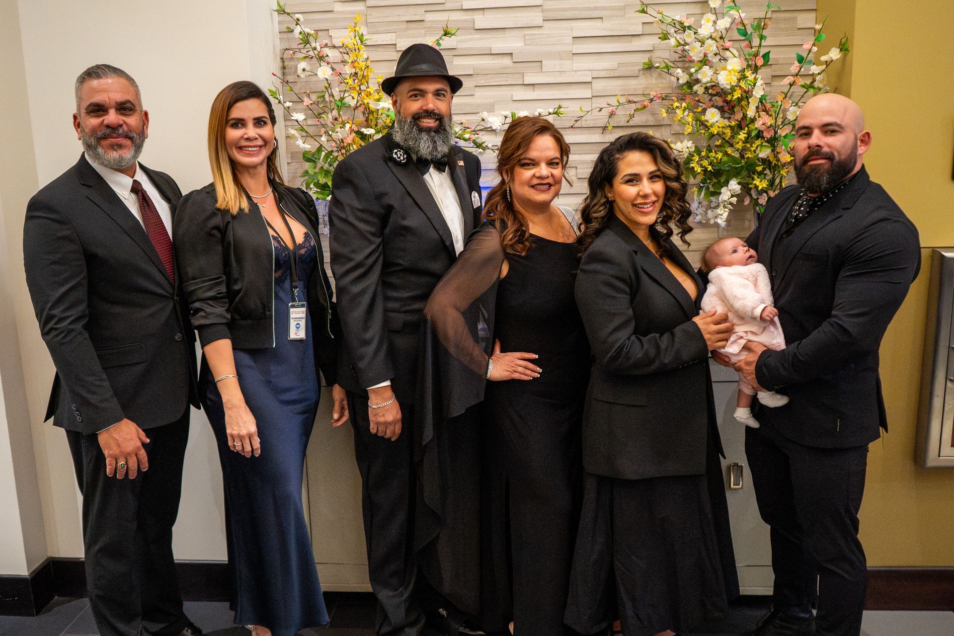 Six people posing indoors in formal attire, one holding a baby, in front of a floral backdrop