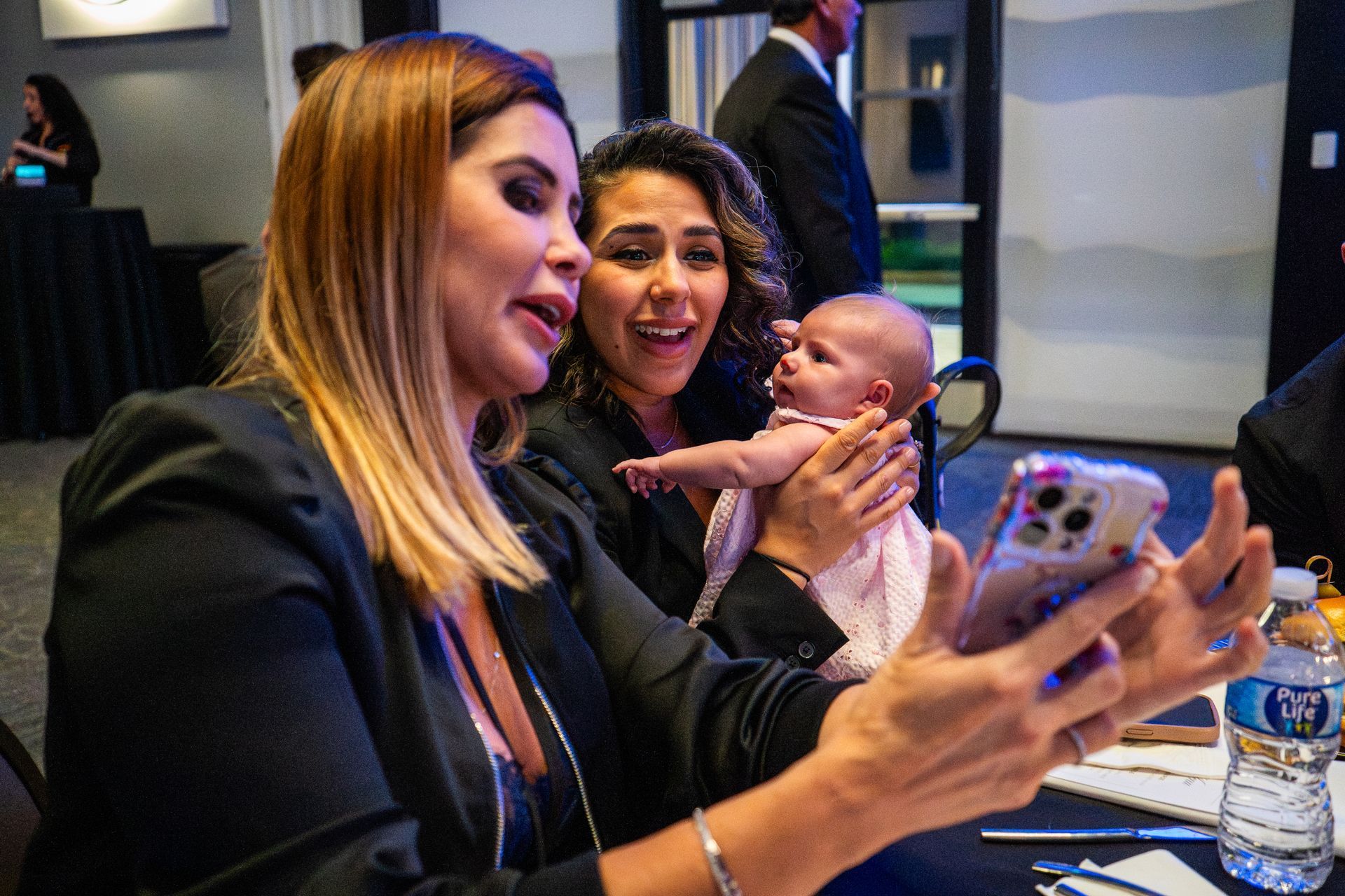 Two women smiling and holding a baby at a table, with one taking a selfie on a phone