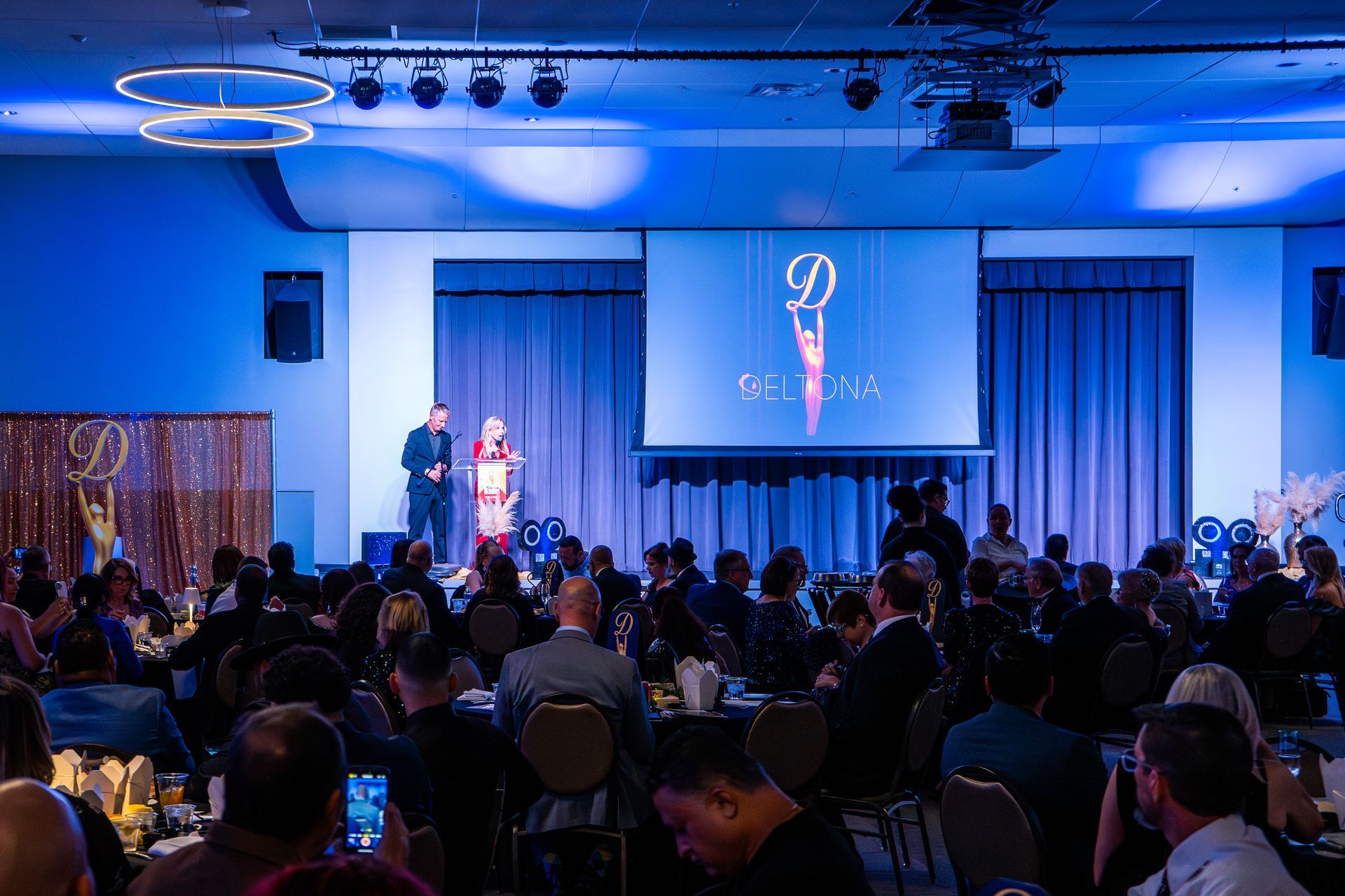 Audience watches a speaker on a blue-lit stage in a crowded conference hall.