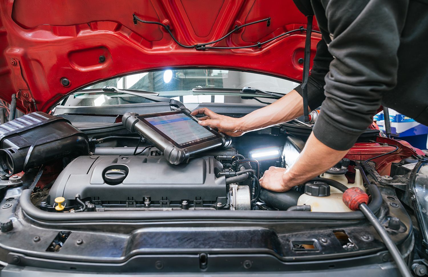 Mechanic using a diagnostic tablet under a red car’s hood in an auto shop