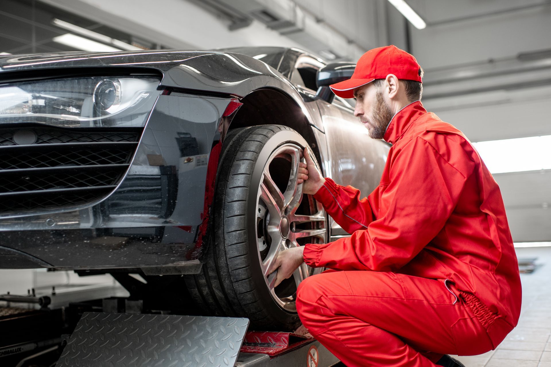 Mechanic in red uniform kneeling beside a black car, inspecting the front wheel in a garage.