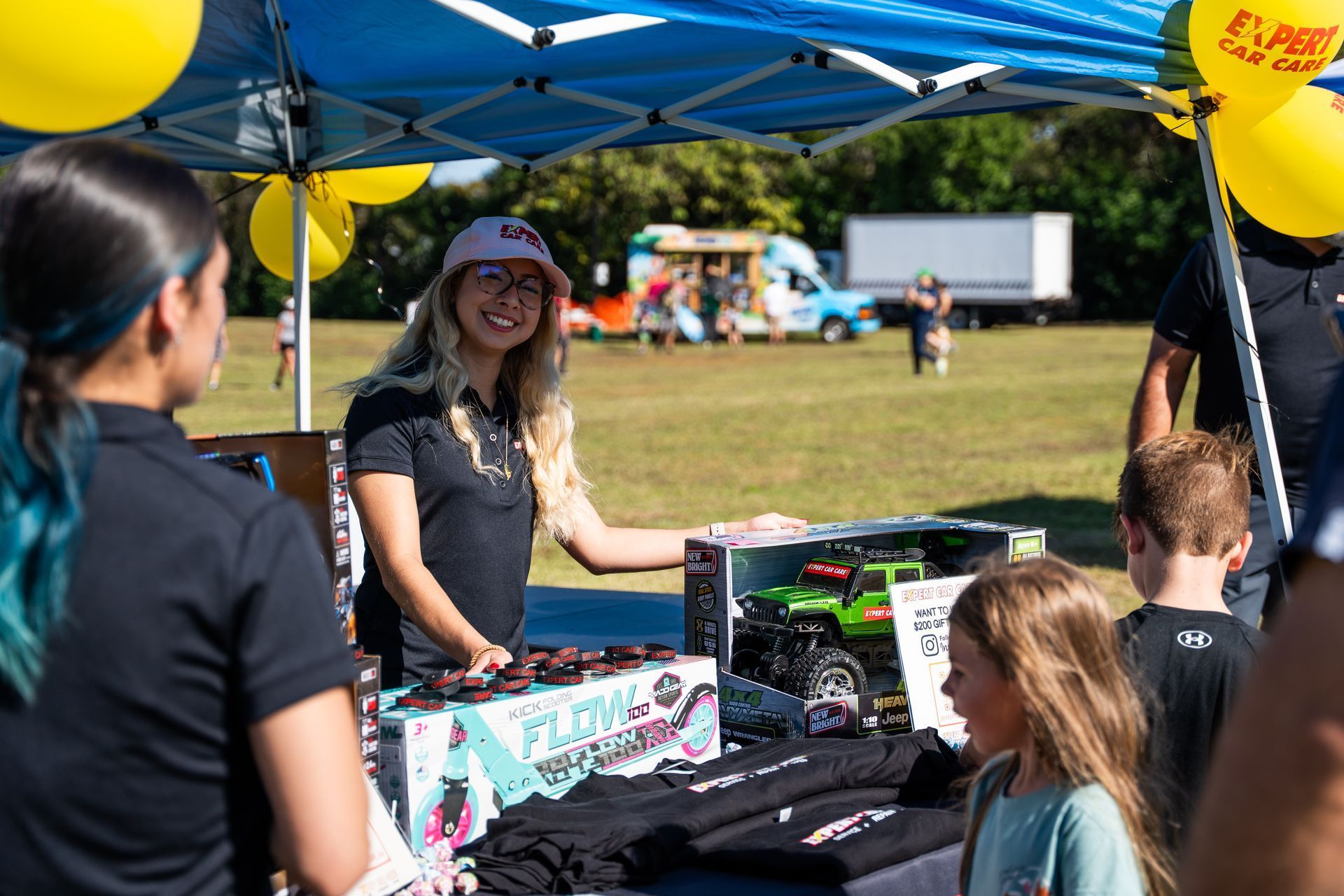 Vendor smiling under blue canopy at an outdoor fair, with children browsing colorful items on a table