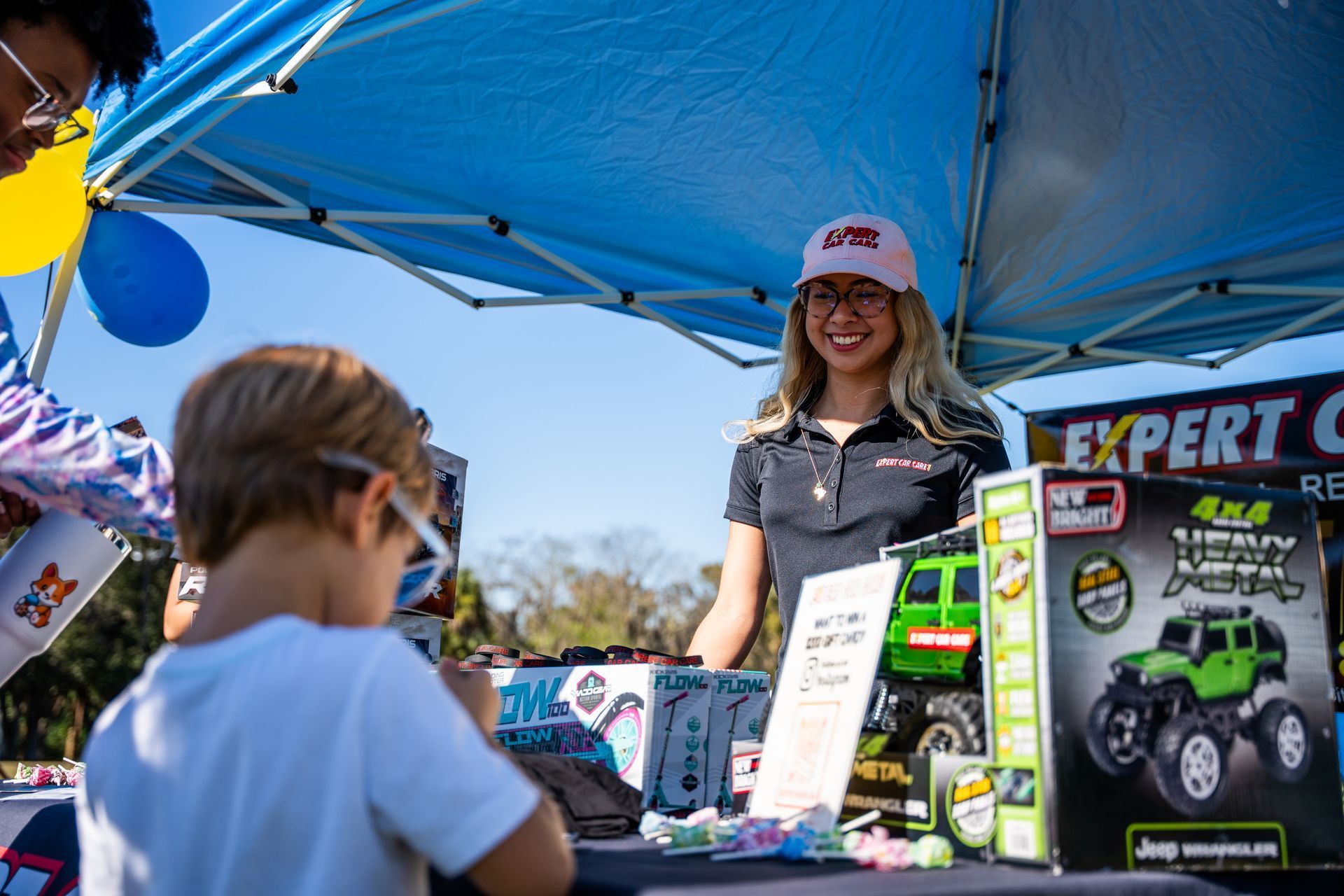 Outdoor booth under blue canopy with promotional toy boxes and a vendor speaking to a child