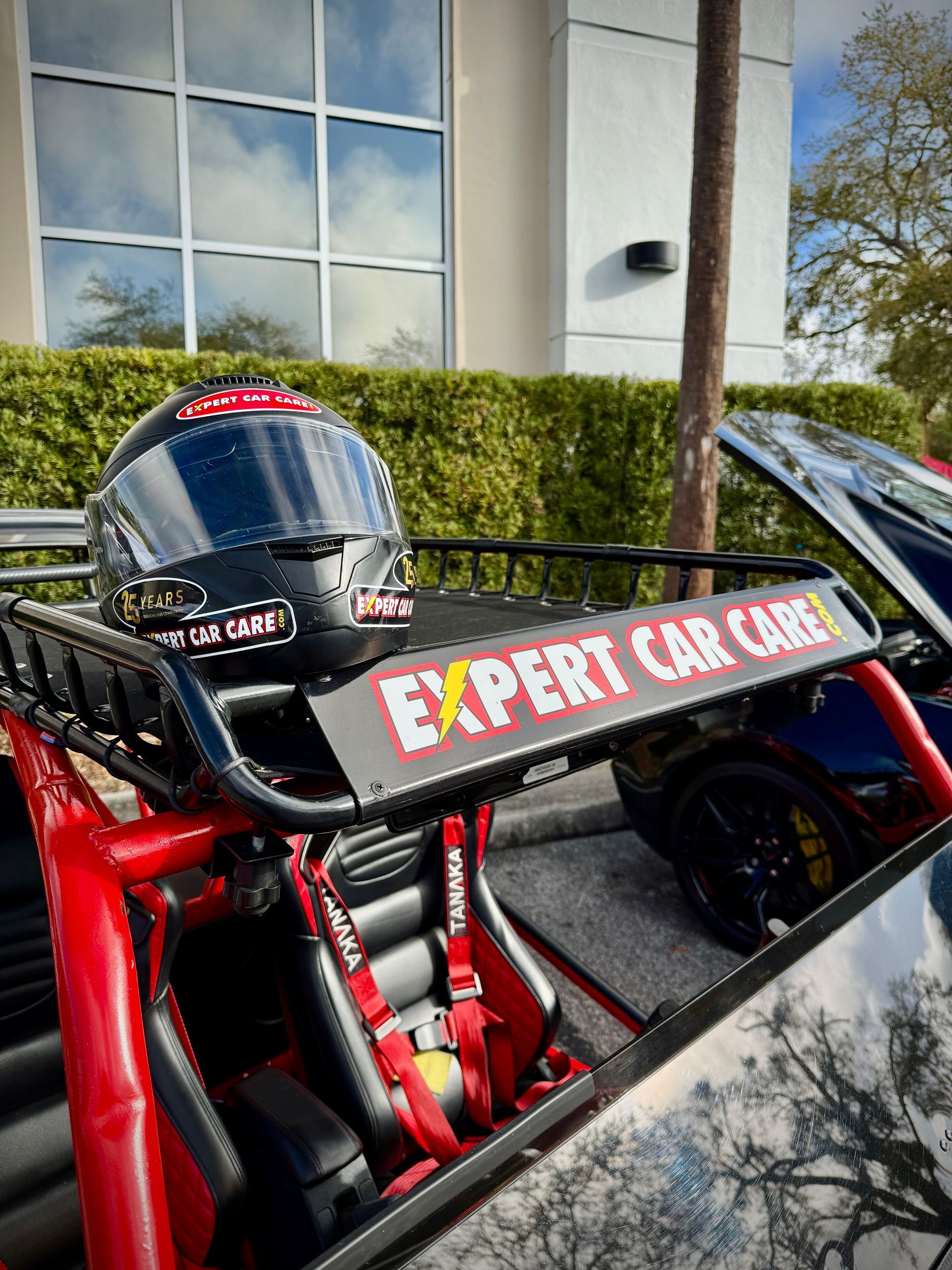 Red open-wheel race car with “EXPERT” branding parked beside a building and hedge