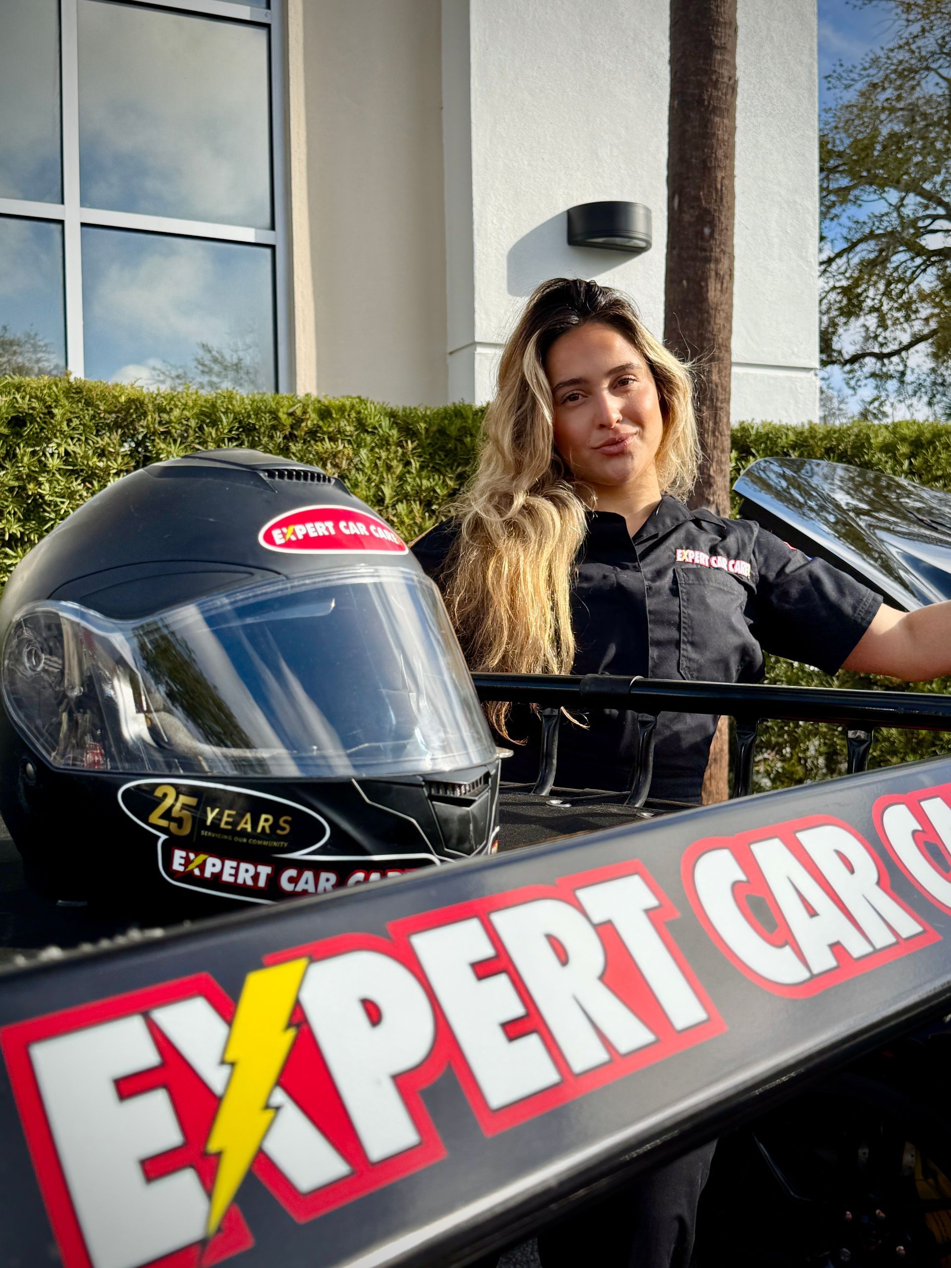 Woman beside race car helmet and banner at outdoor car show, standing by a building