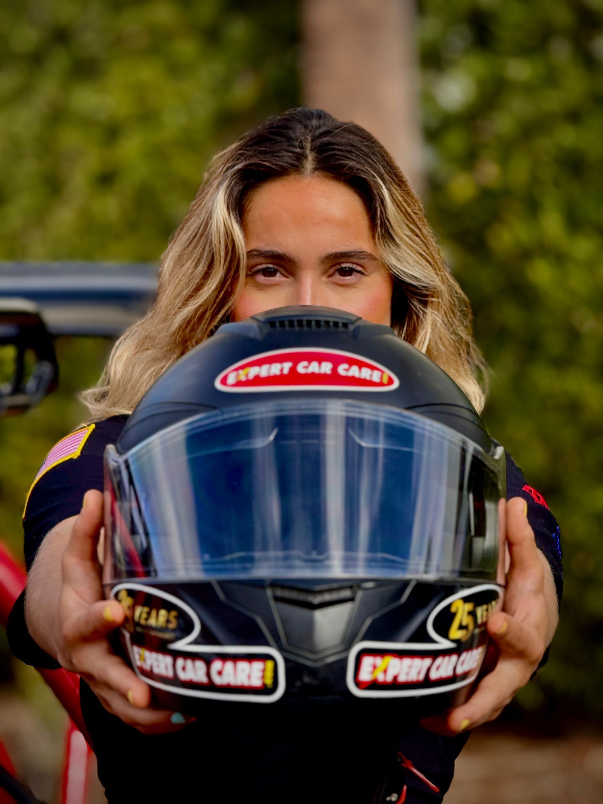 Person holding out a black racing helmet with red-and-white sponsor logos outdoors