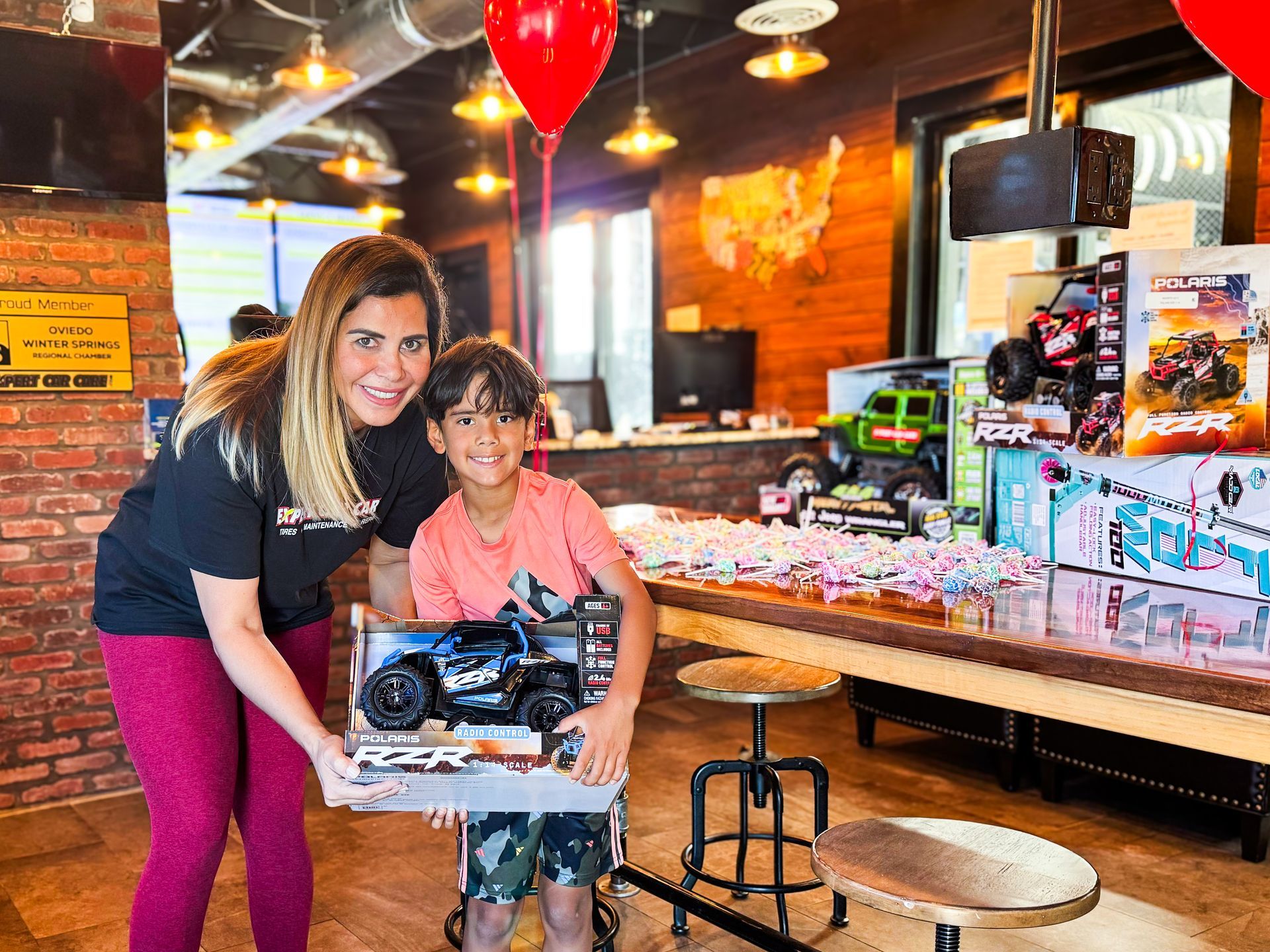 Woman and child posing with a toy car in a restaurant, with a red balloon and counter in the background