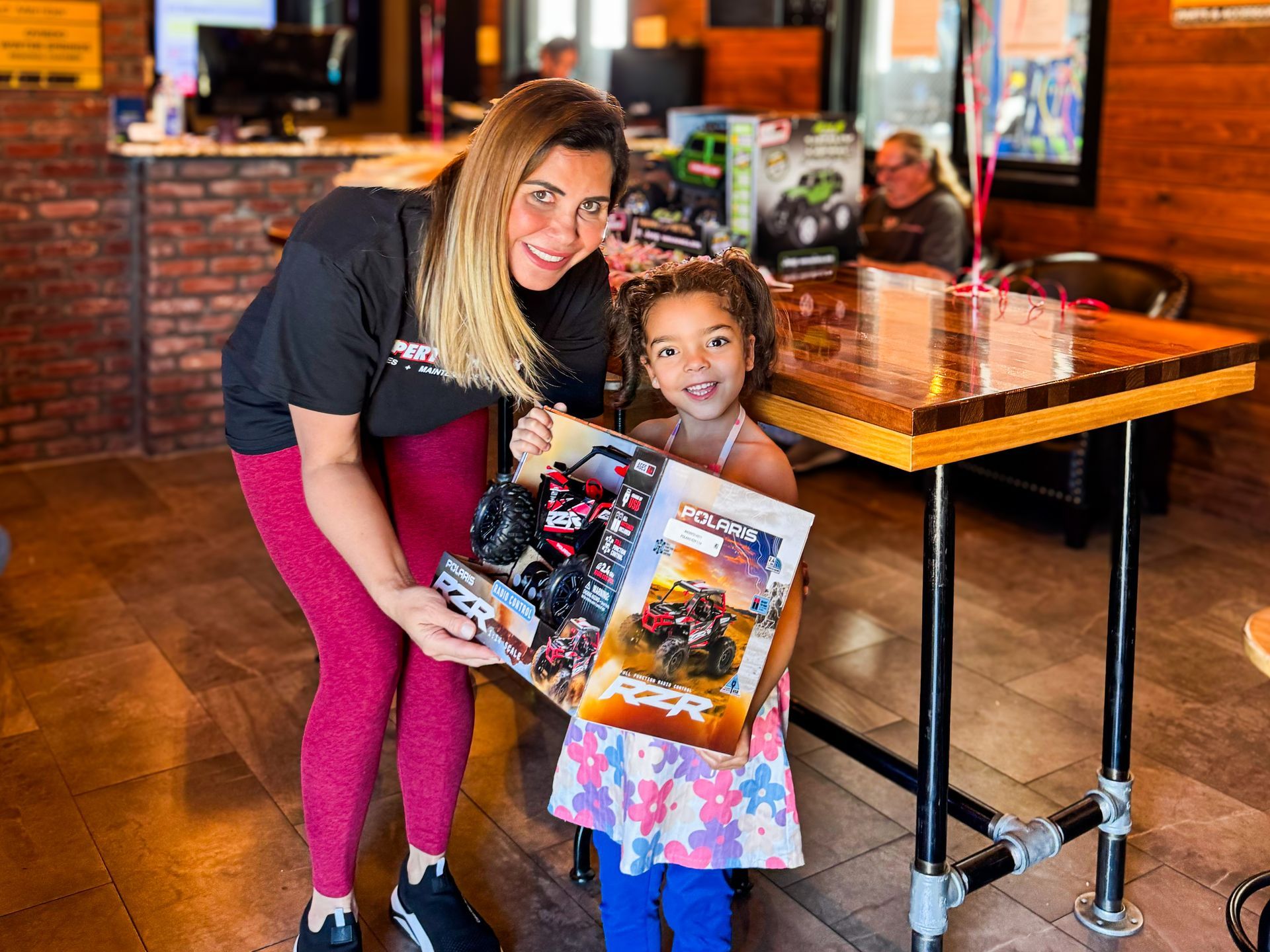 Woman and child smiling in a restaurant, holding colorful toy boxes near a wooden table.