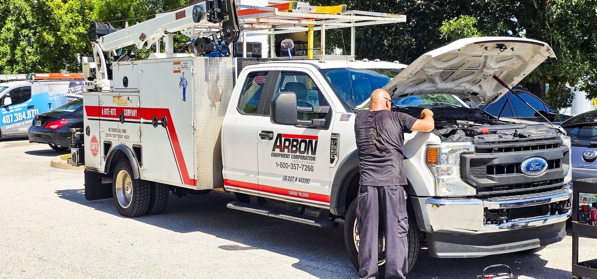Tow truck with hood open, a worker at the front, parked on a sunny street beside a blue car.