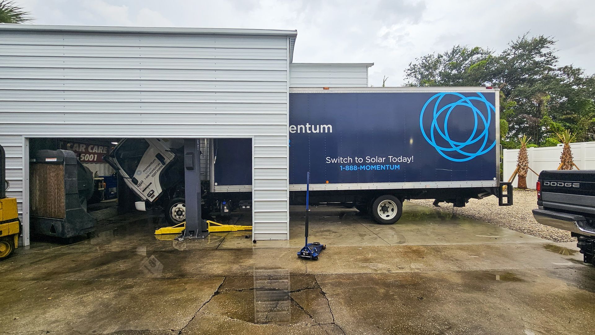 Blue and black truck parked beside a white industrial building on a wet concrete lot