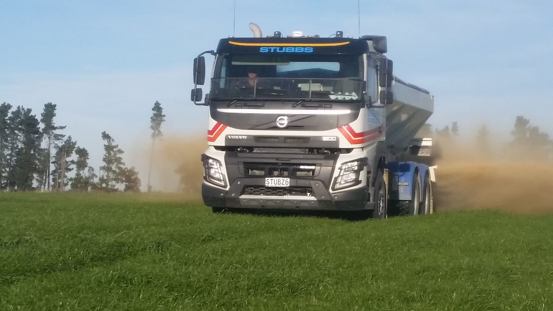 White fertilizer truck drives through a green field, kicking up dust under a clear blue sky.