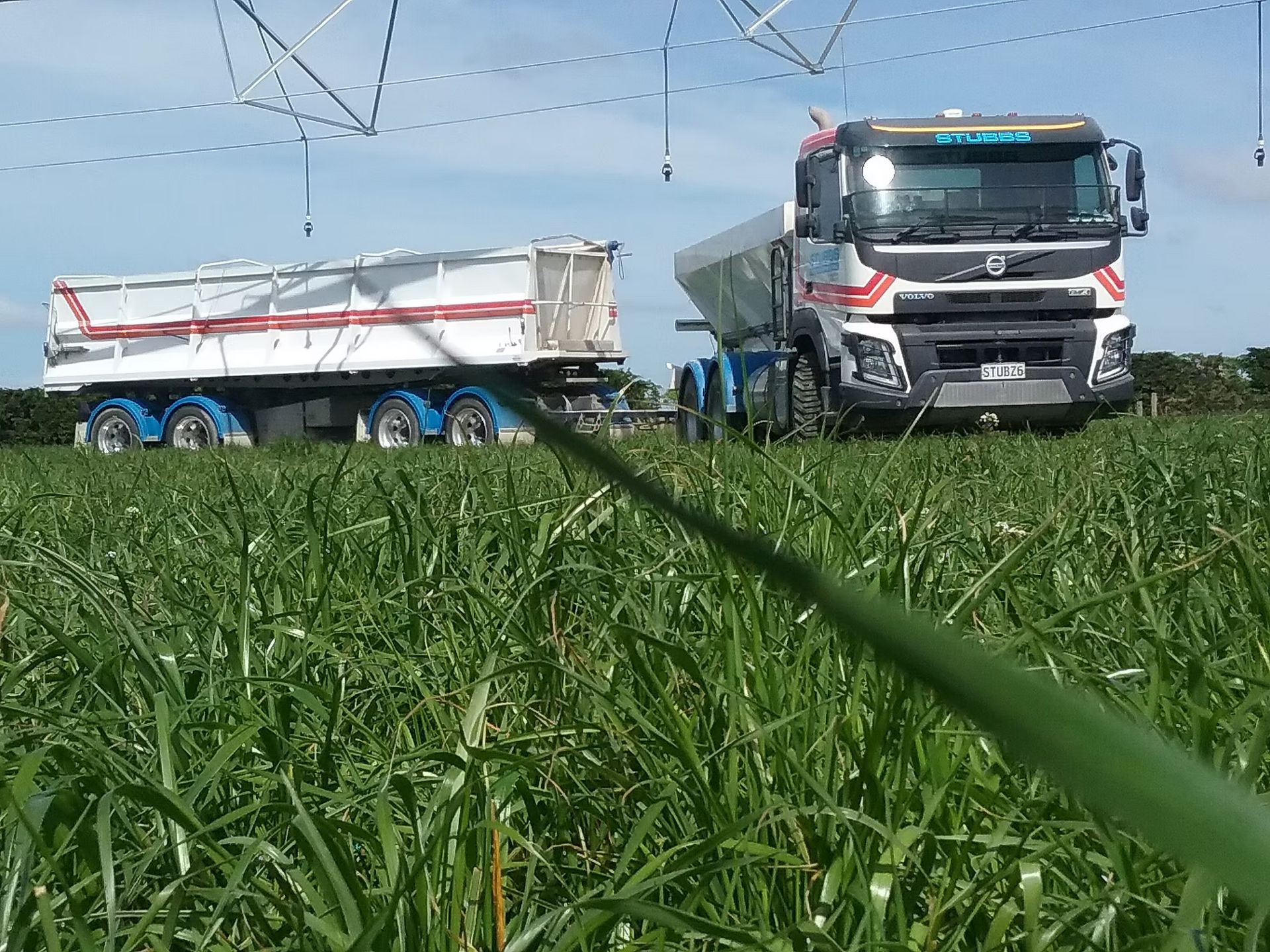 Two white trucks on a grassy field under power lines, one towing a white trailer.