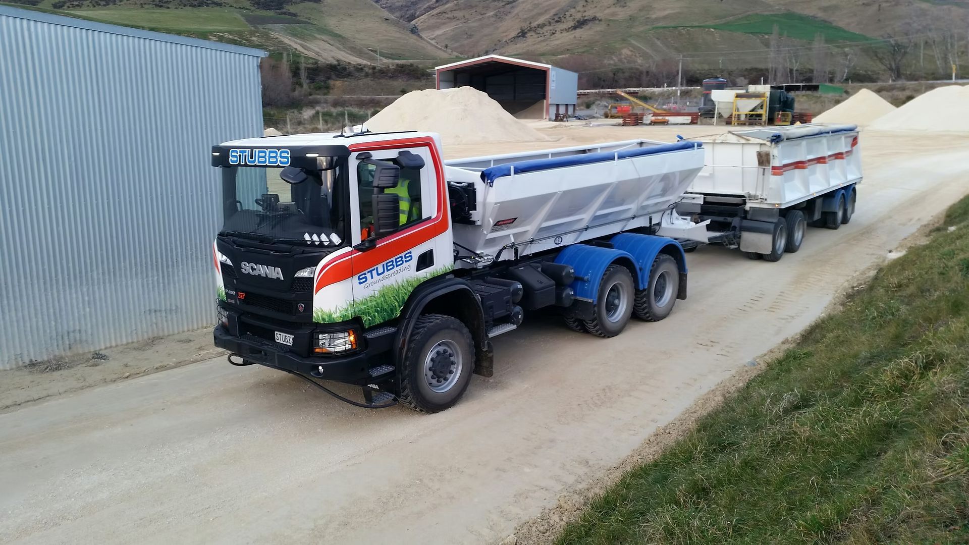 Large blue dump truck hauling a trailer on a dirt road in a rural area