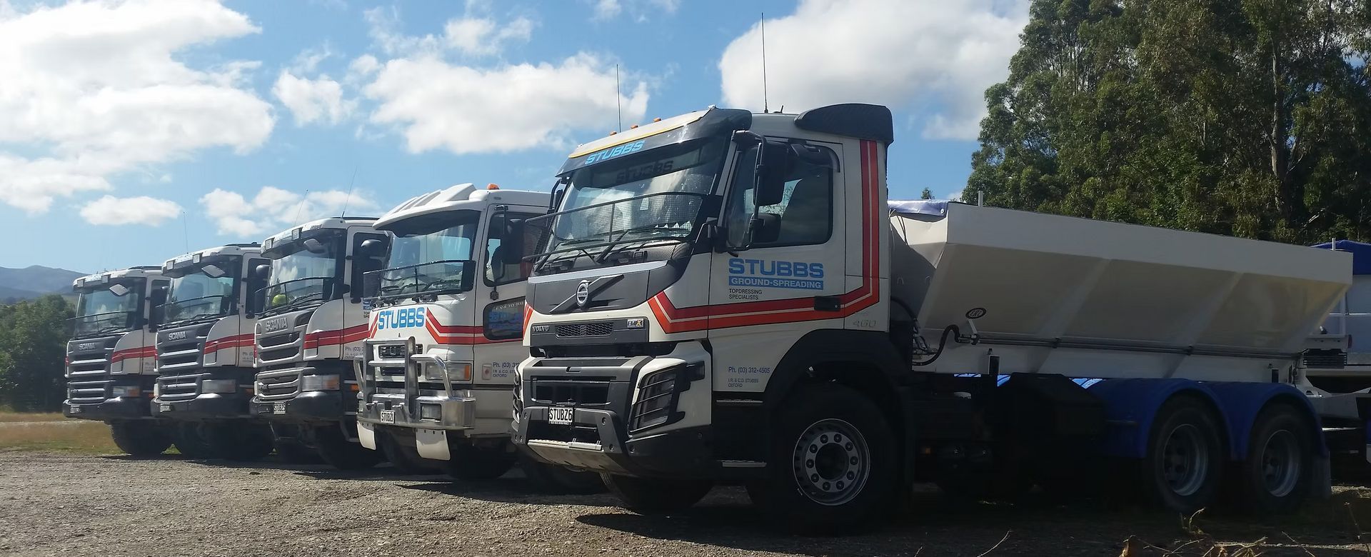 Line of white dump trucks parked in a row on a gravel lot under a blue sky